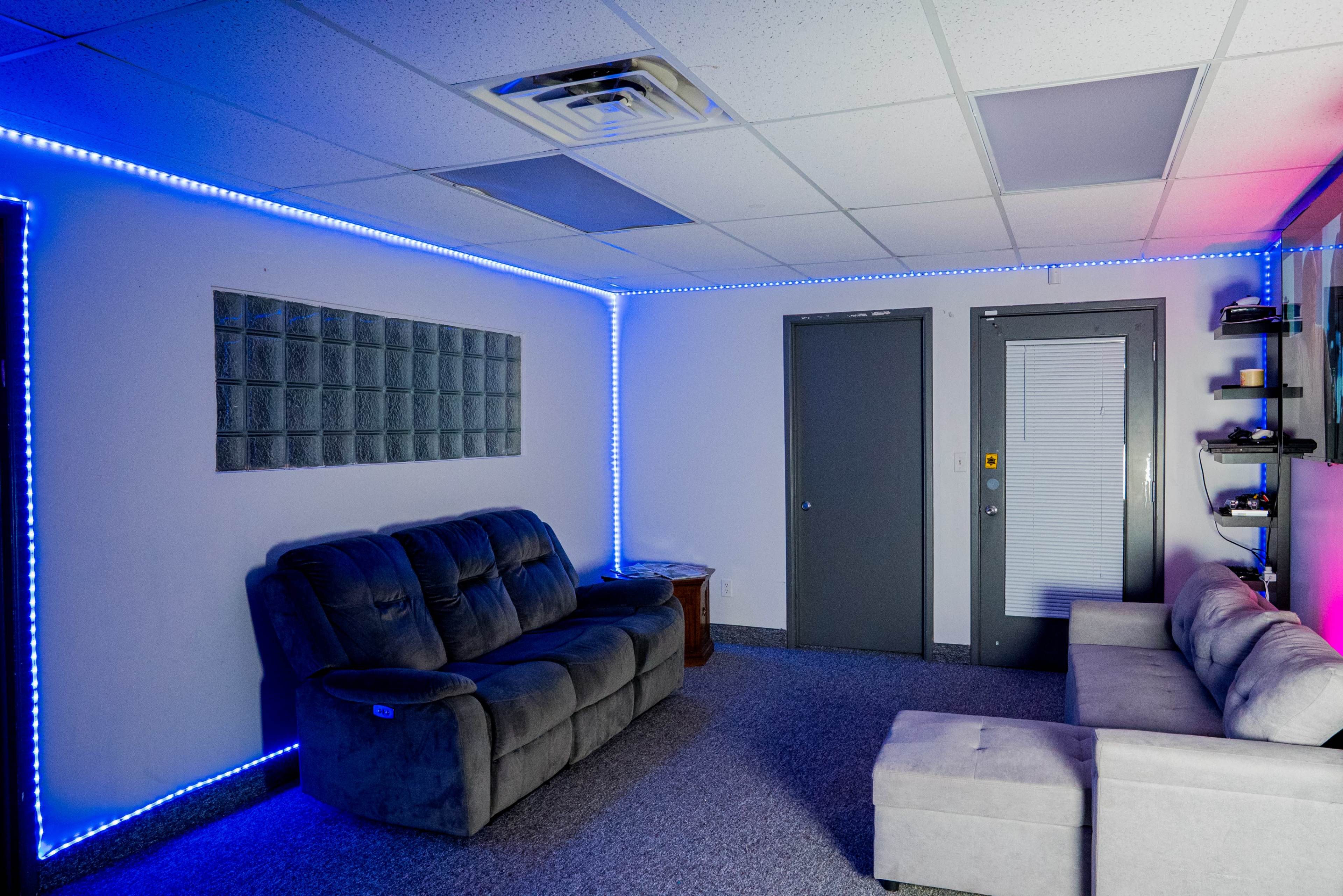 A room features two sofas, one gray recliner and one light-colored couch, with blue LED lights outlining the walls and a textured glass block panel on one side.
