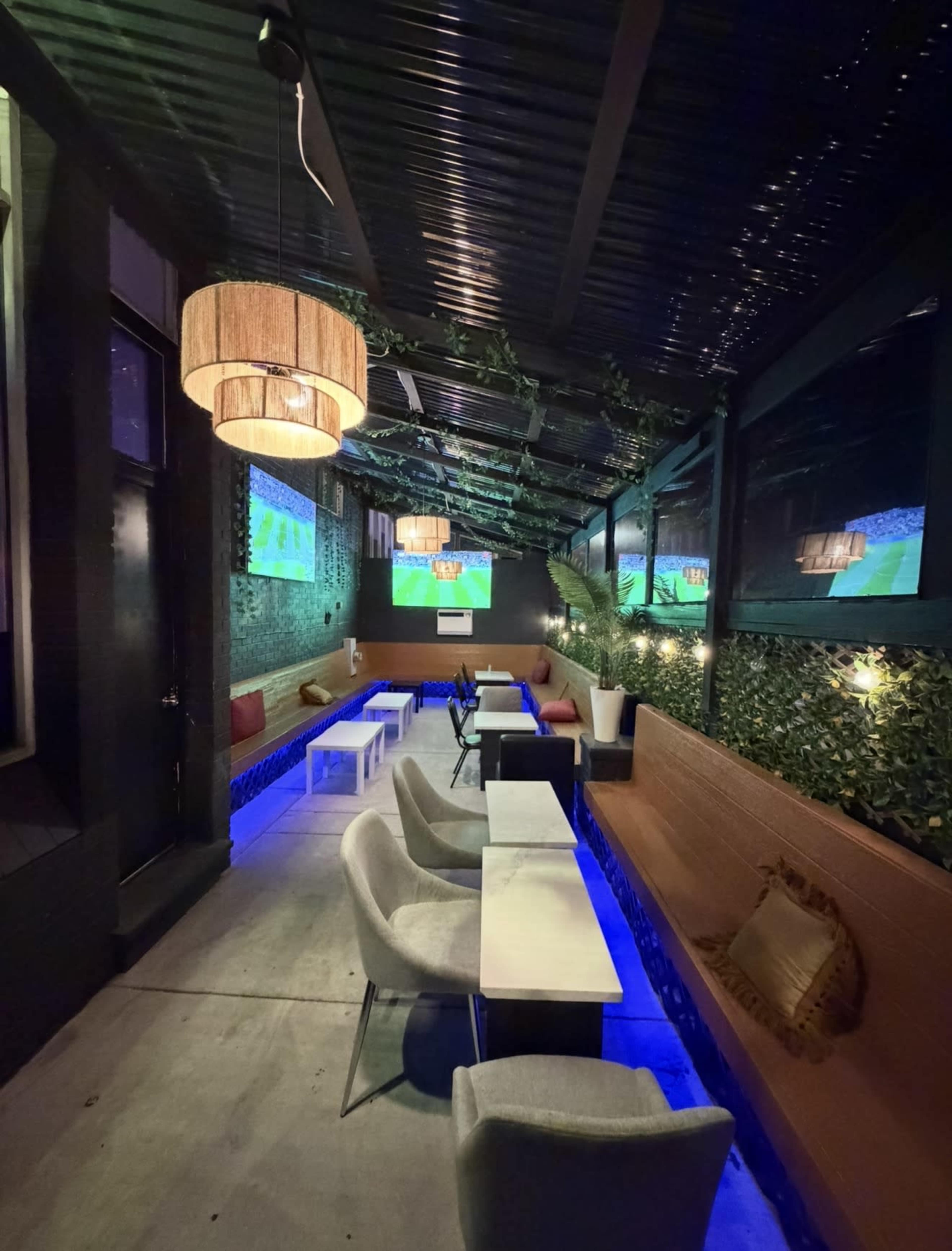 The image shows a partially enclosed lounge area with modern furniture, ambient lighting, and a large screen displaying sports, surrounded by greenery.