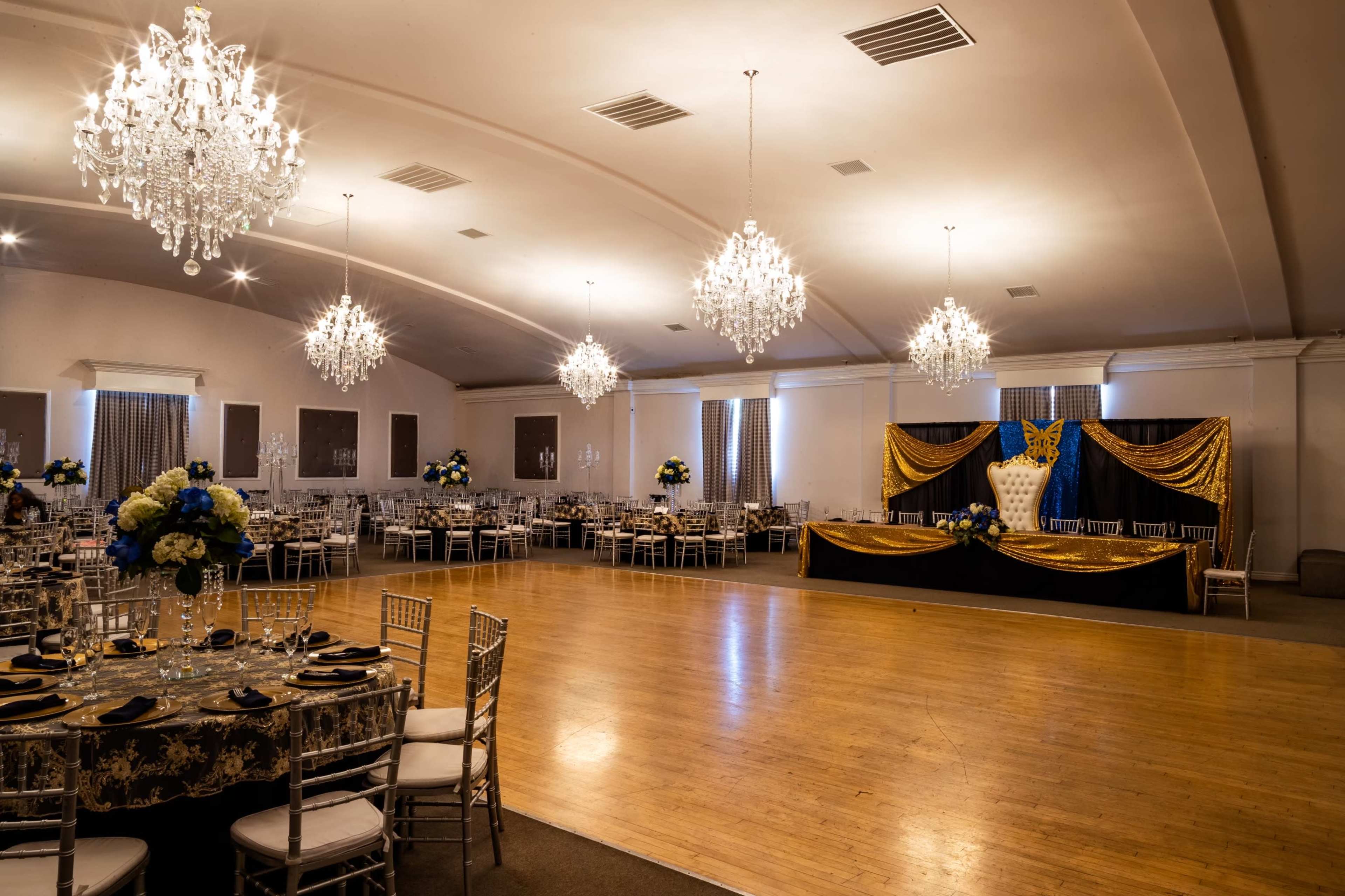 A large event space features round tables with elegant table settings, crystal chandeliers, and a decorated stage with a cake backdrop.