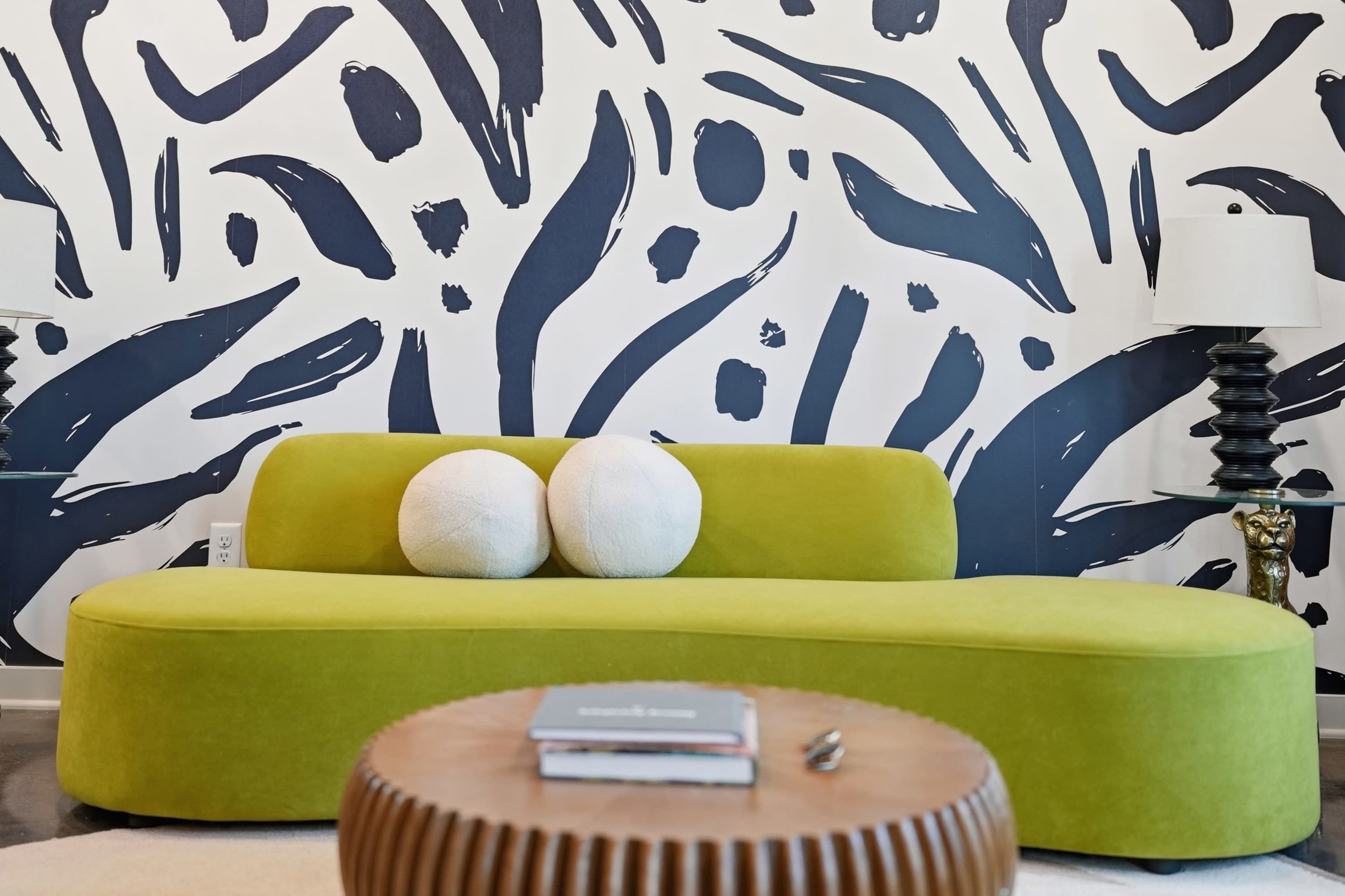 The image features a modern curved green sofa adorned with two white round cushions, set against a striking black and white patterned wall, with a wooden coffee table in the foreground.