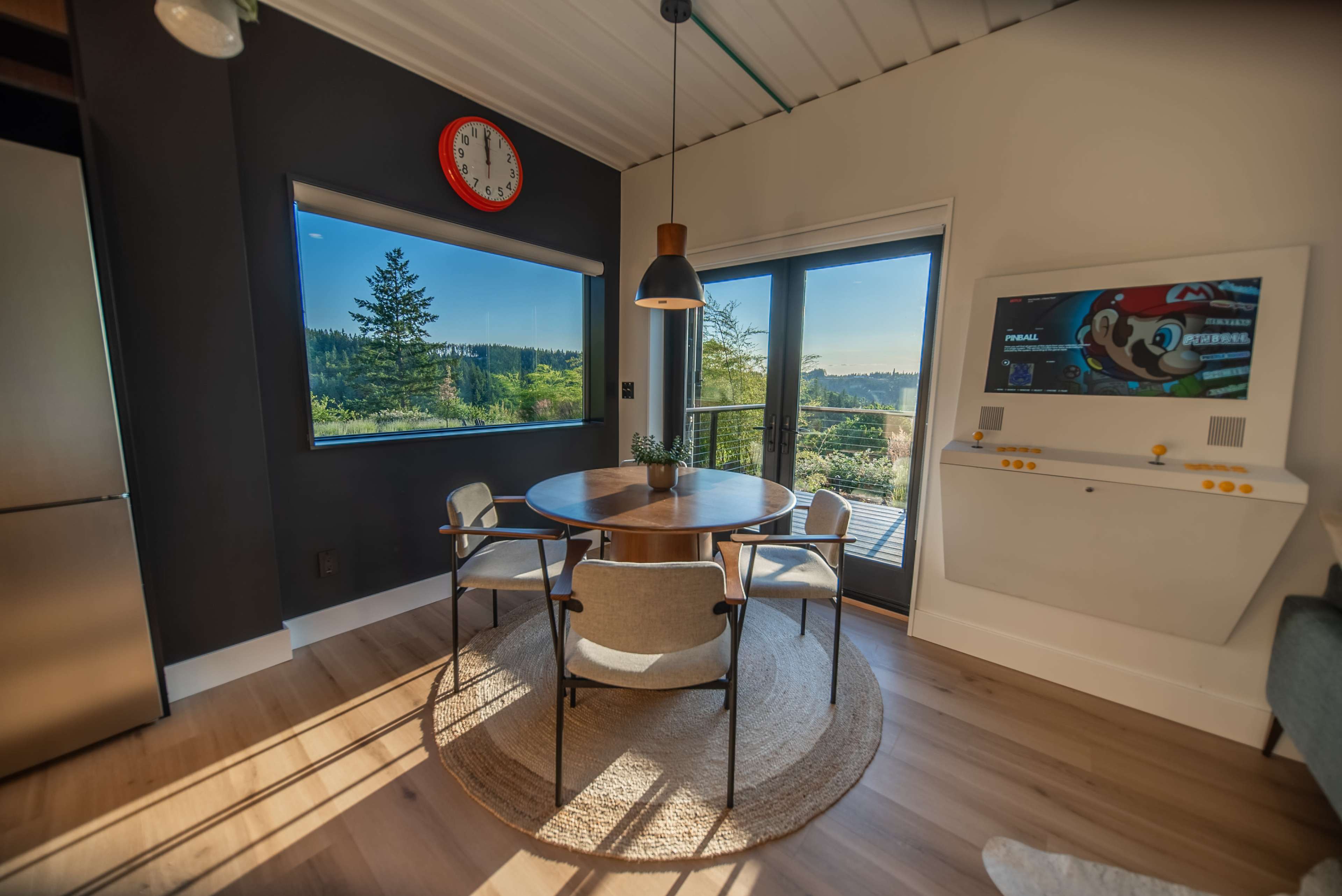 A modern dining area features a circular table with four chairs, large windows showcasing a scenic view, and a wall-mounted TV beside a cheerful clock.