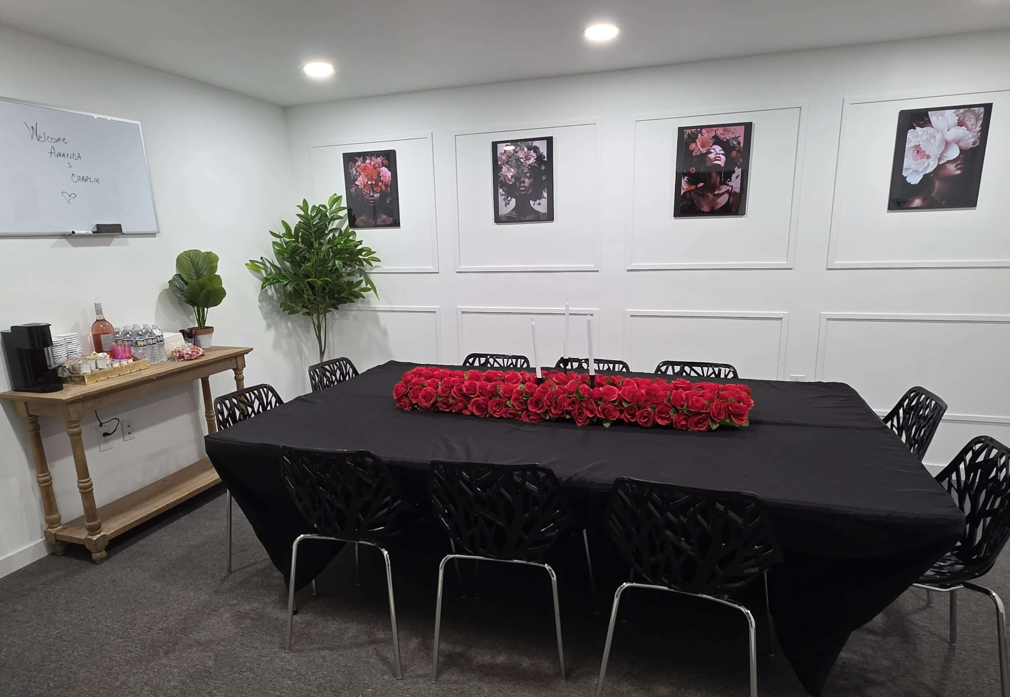 The image shows a well-decorated meeting room with a large table covered in a black tablecloth, surrounded by black chairs and adorned with a row of red roses in the center, complemented by floral artwork on the walls.
