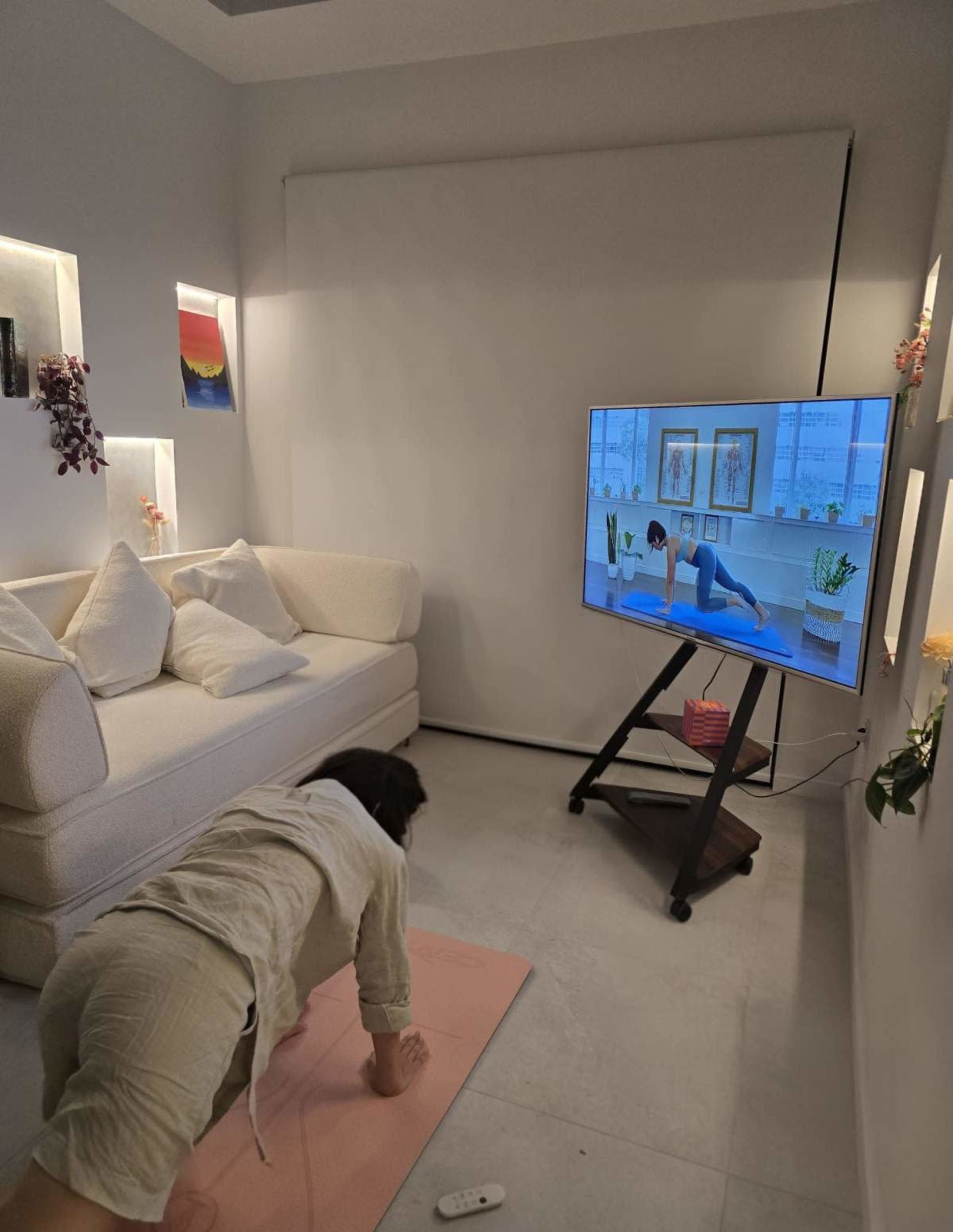 A person performs an exercise on a pink mat in a modern living room while following a workout video displayed on a television.