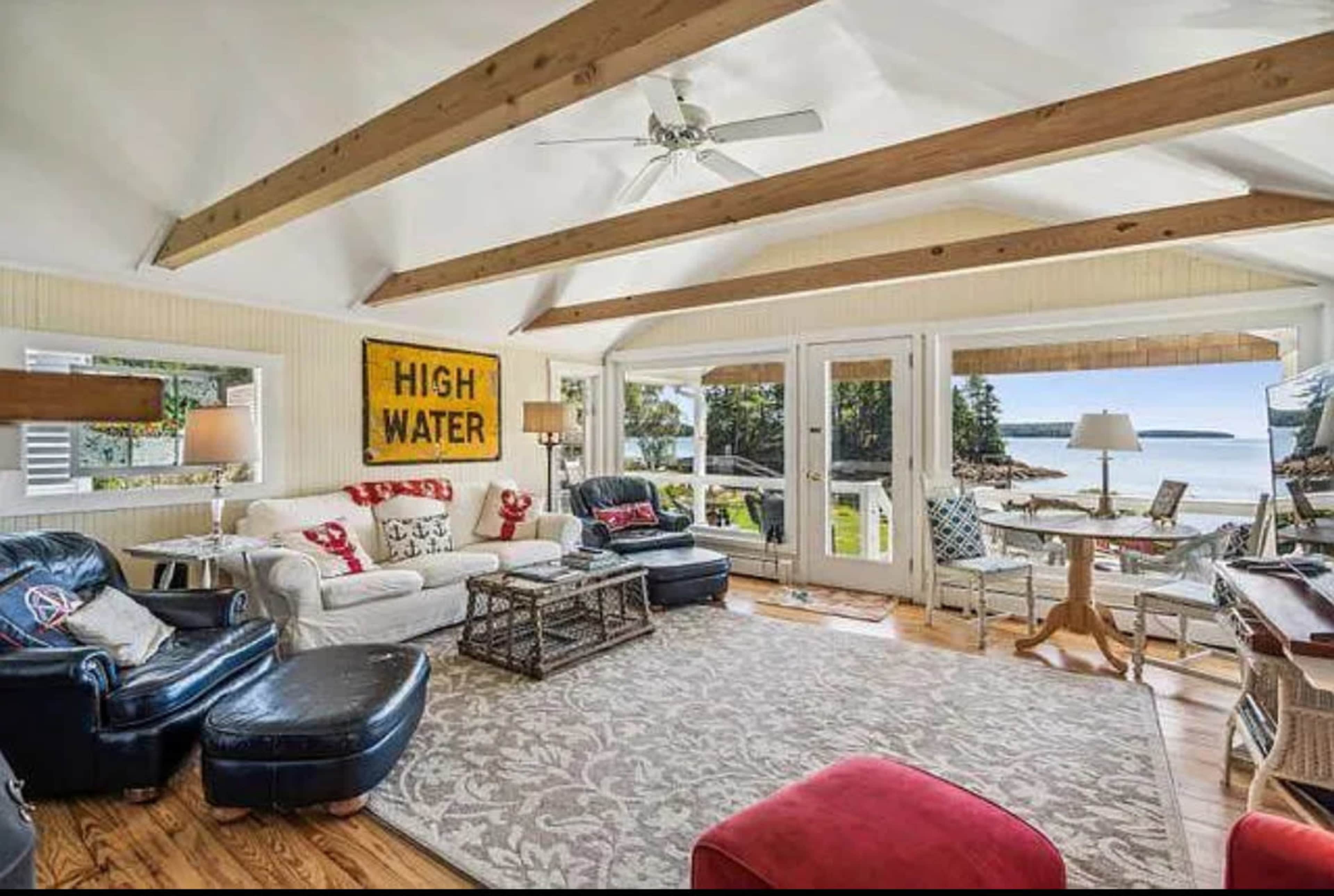 A living room features a mix of comfortable seating, a rug, and large windows that offer a view of the water.