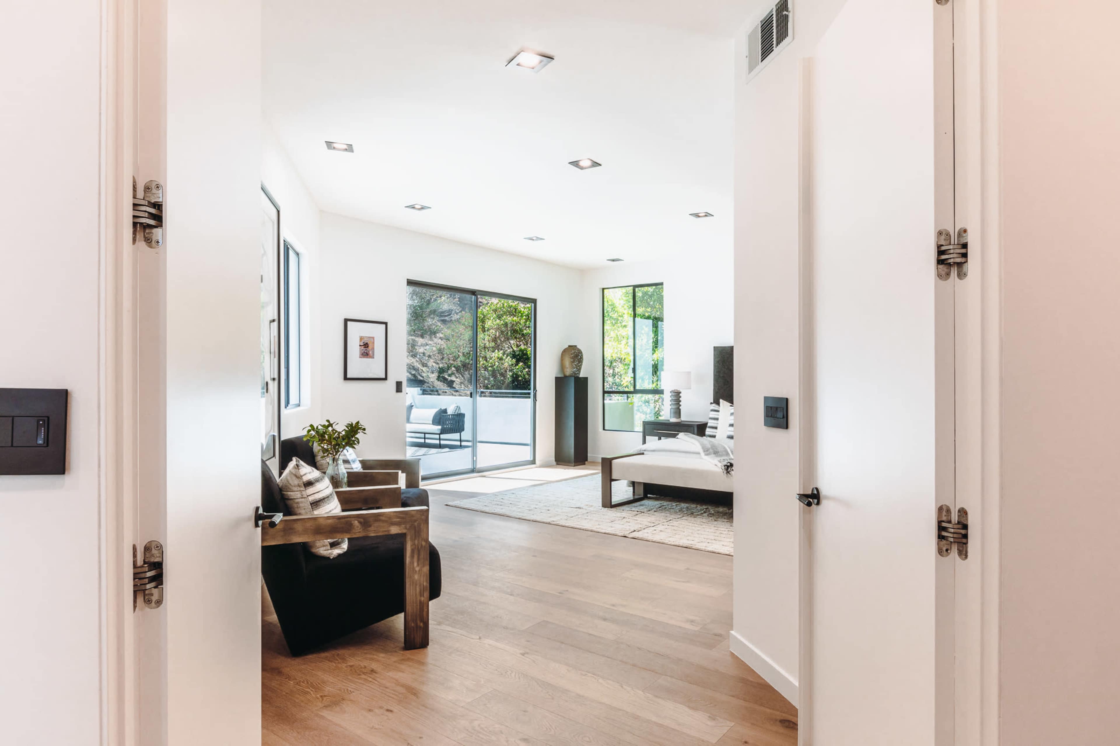An open doorway leading into a modern living room with large windows, hardwood floors, and minimalistic decor.
