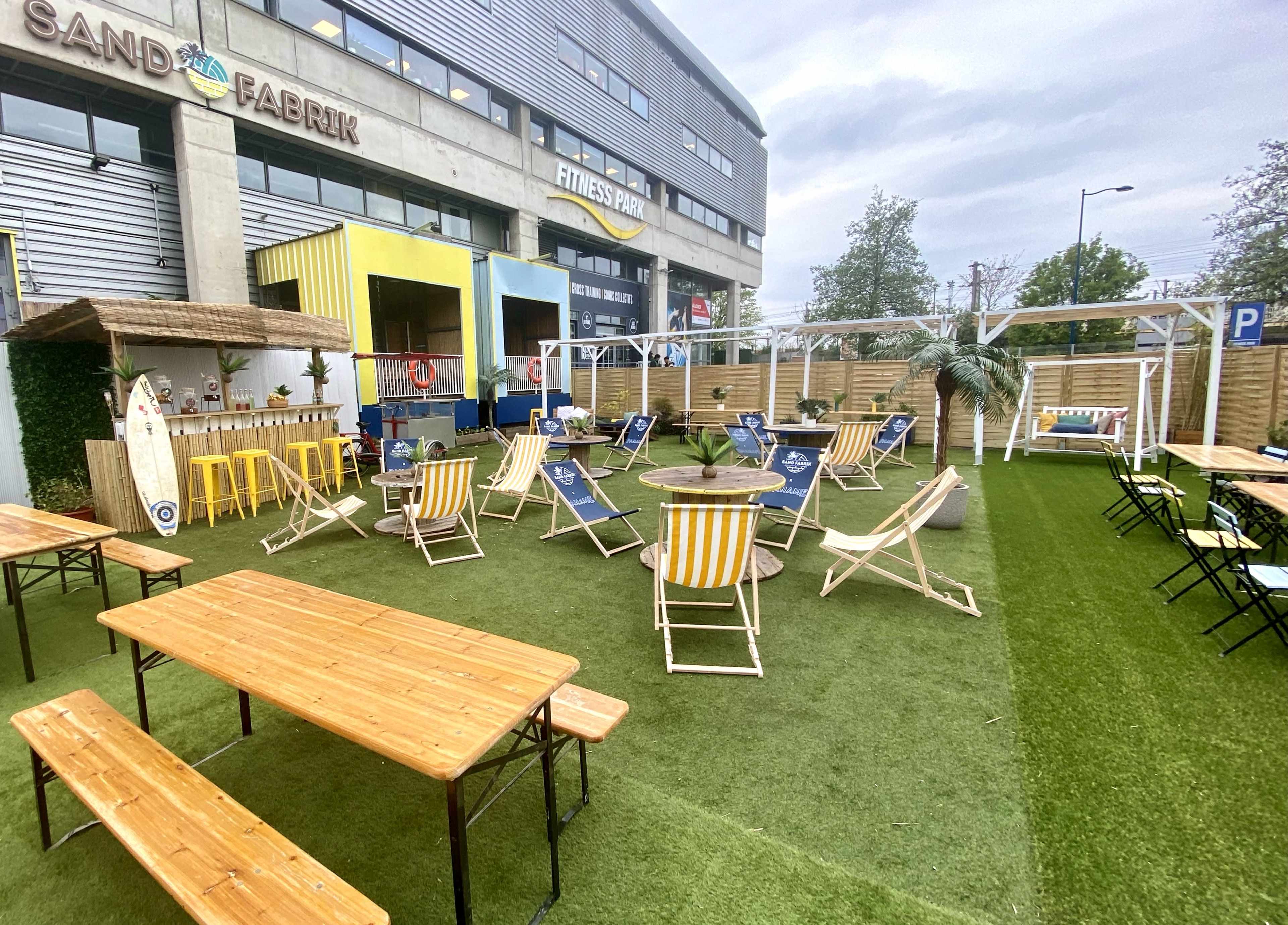 An outdoor seating area features wooden picnic tables, lounge chairs, and greenery, set against a backdrop of a modern building.