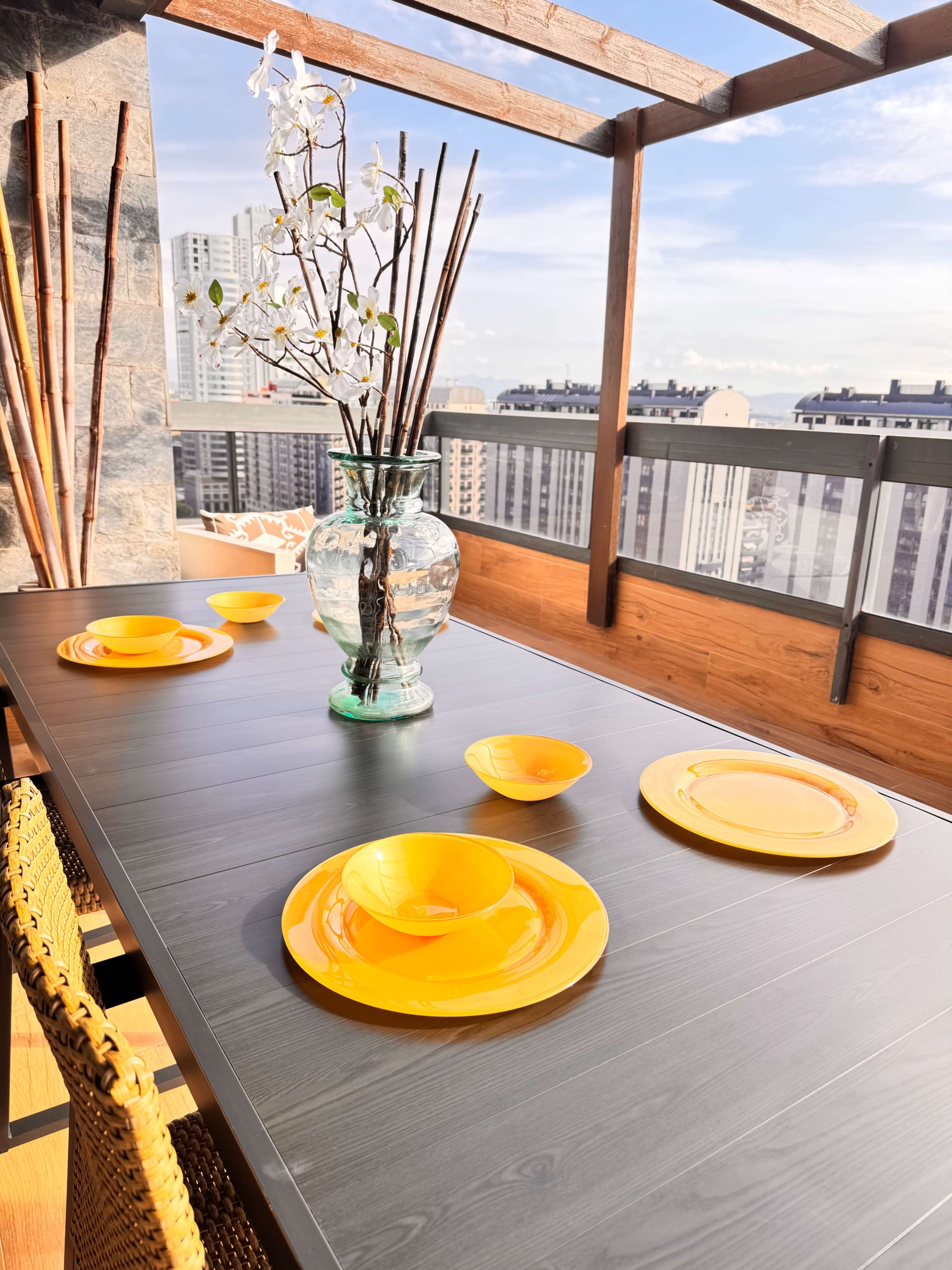 A dining table with yellow plates and bowls is set next to a vase of flowers, overlooking a city skyline from a balcony.