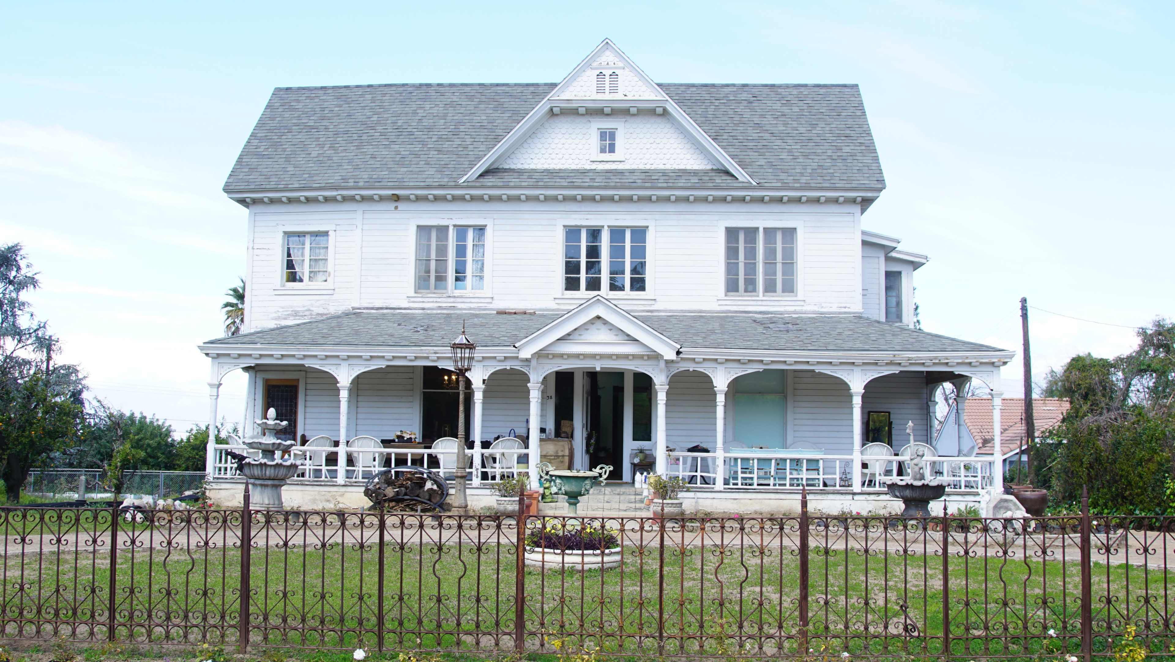 A large, two-story white house with a decorative porch and a well-maintained yard is surrounded by a wrought-iron fence.