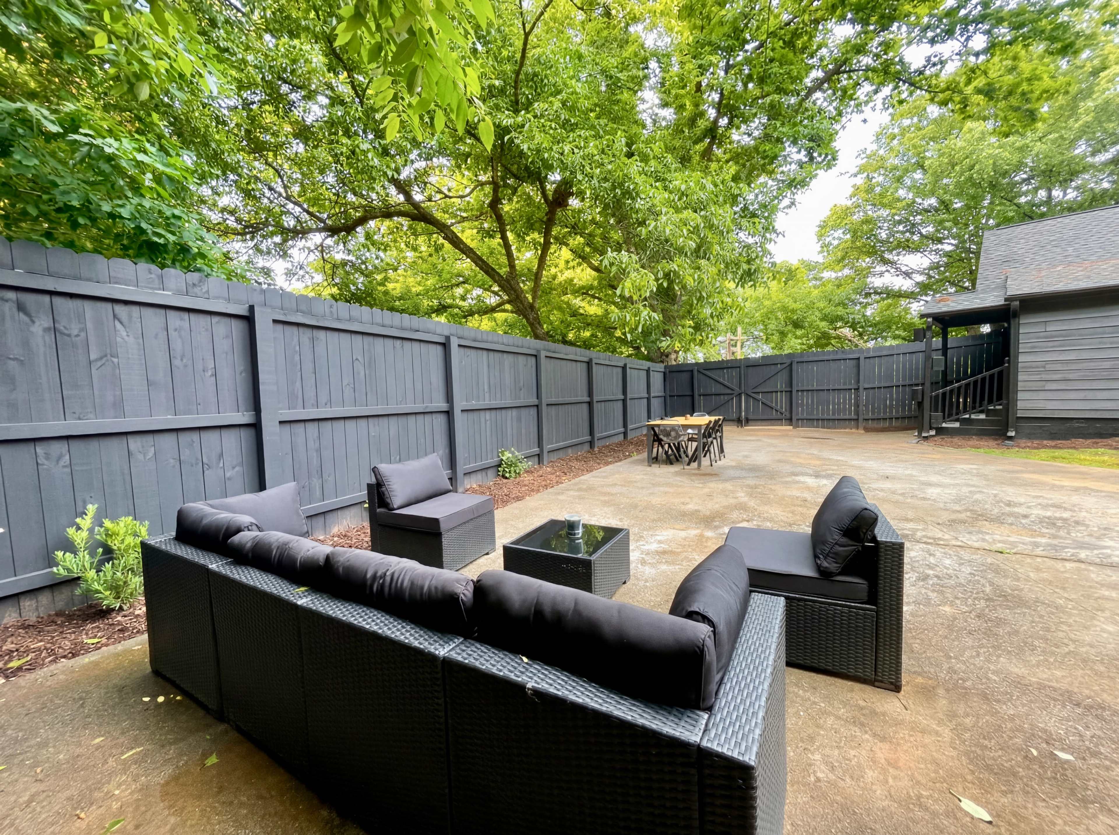 The image shows a landscaped outdoor patio area with a dark wooden fence, a seating arrangement made of black wicker furniture, and a wooden dining table surrounded by chairs.
