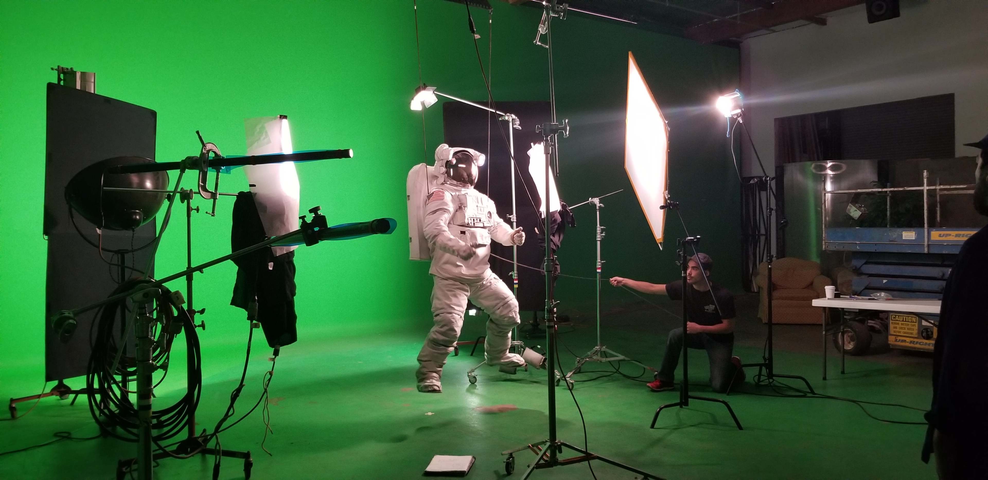 A person in a space suit stands in front of a green screen while filming equipment and lights are positioned around them.