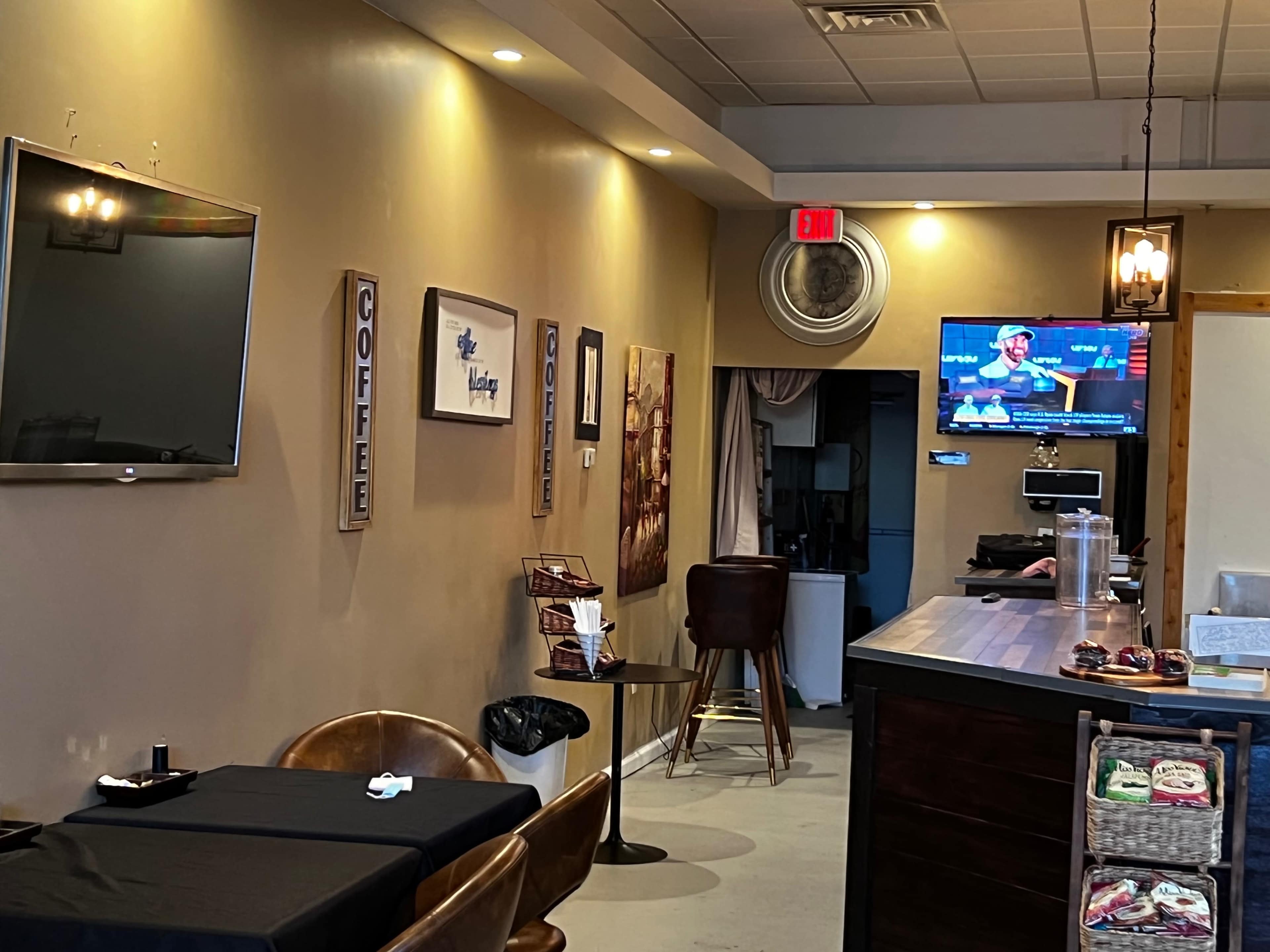 A modern café interior featuring a dark table, brown chairs, a wall-mounted television, and decor on the walls.