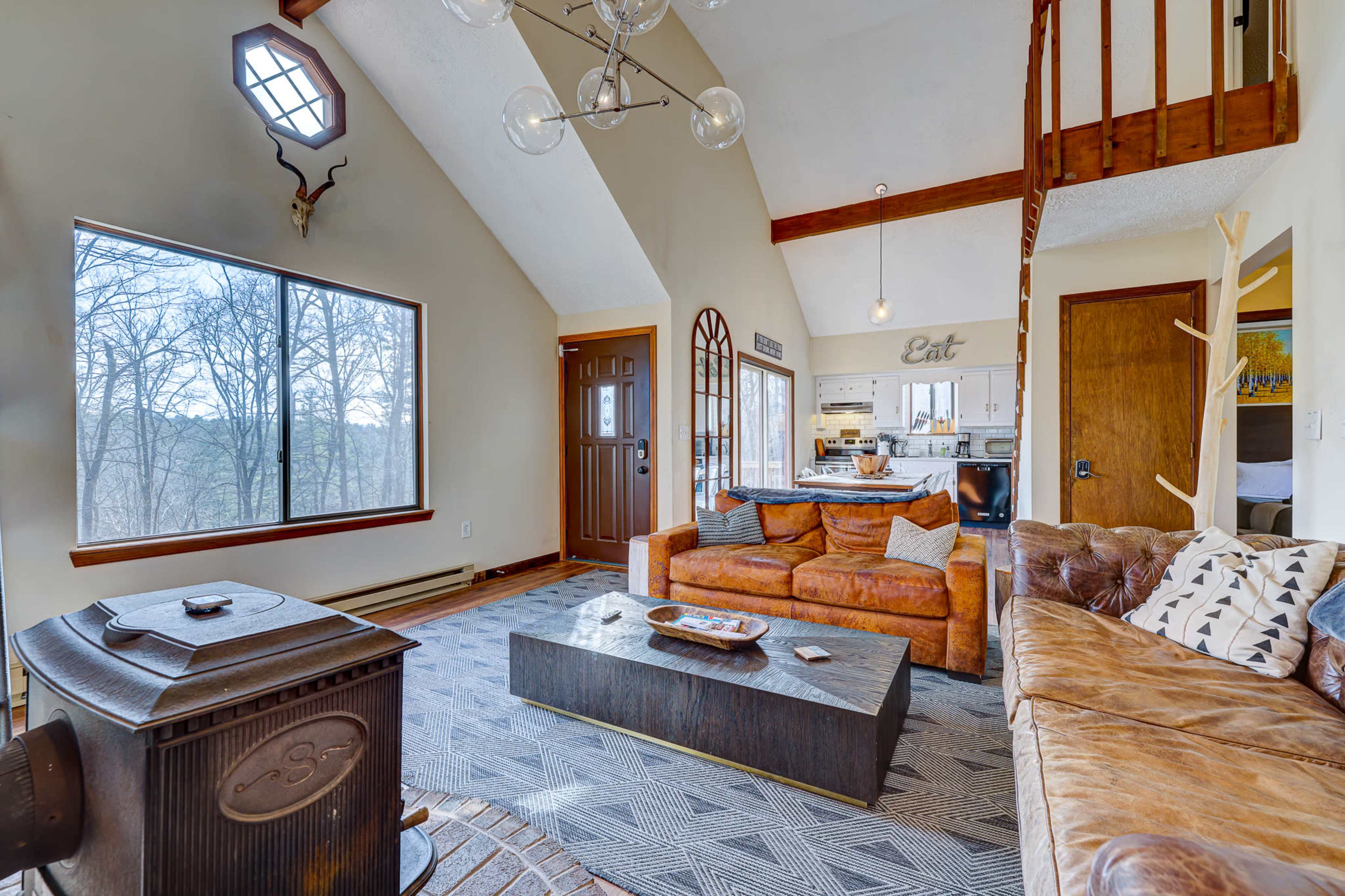 The living room features a combination of a brown leather sofa and an armchair, a coffee table in the center, large windows offering a view of the outdoors, and a stove in the corner.