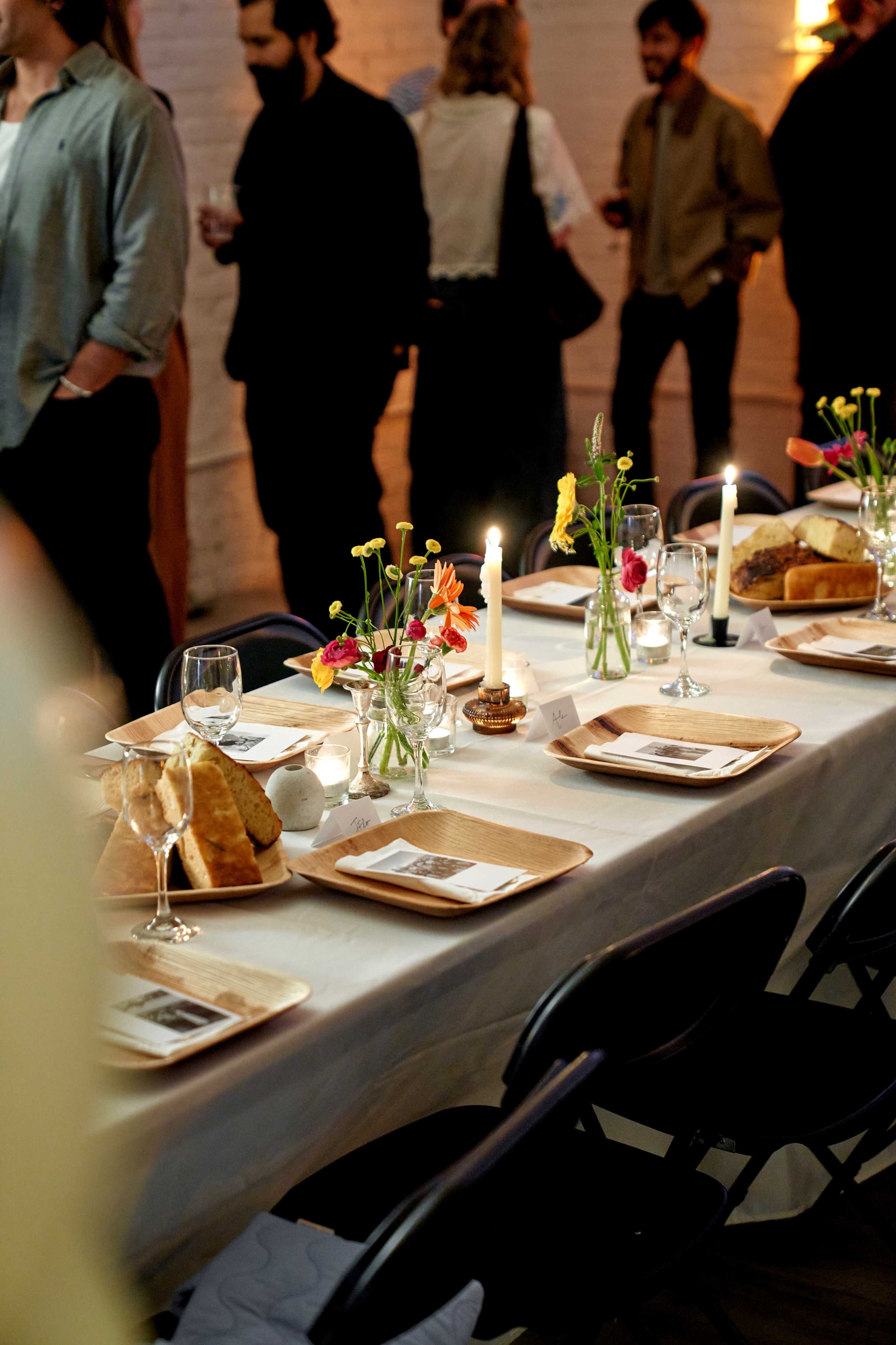 A long dining table is set with plates of food, glassware, and small flower arrangements, while people gather in the background.