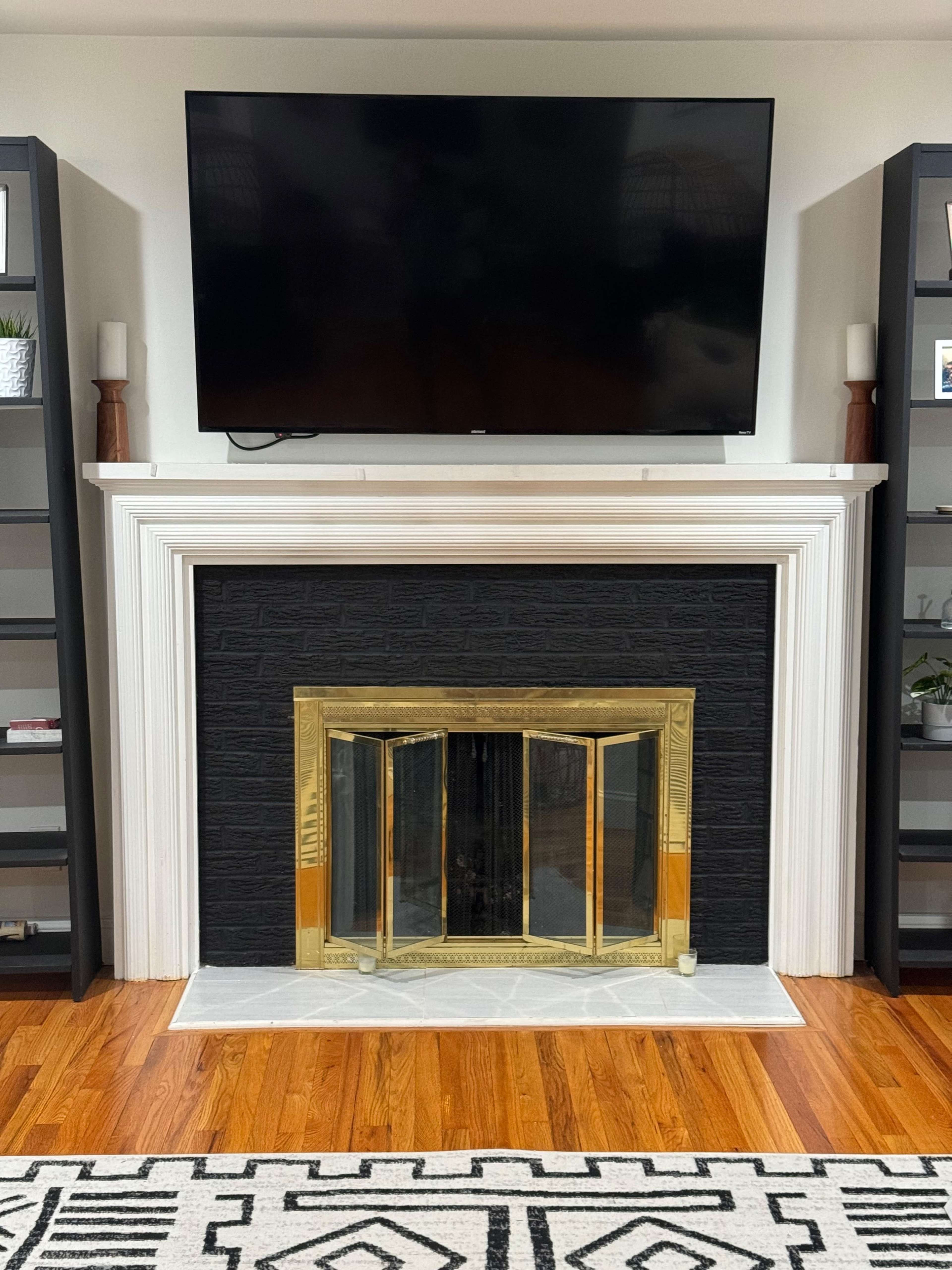 A living room with a black stone fireplace featuring a gold-framed screen, placed beneath a large flat-screen TV, flanked by two dark bookshelves.