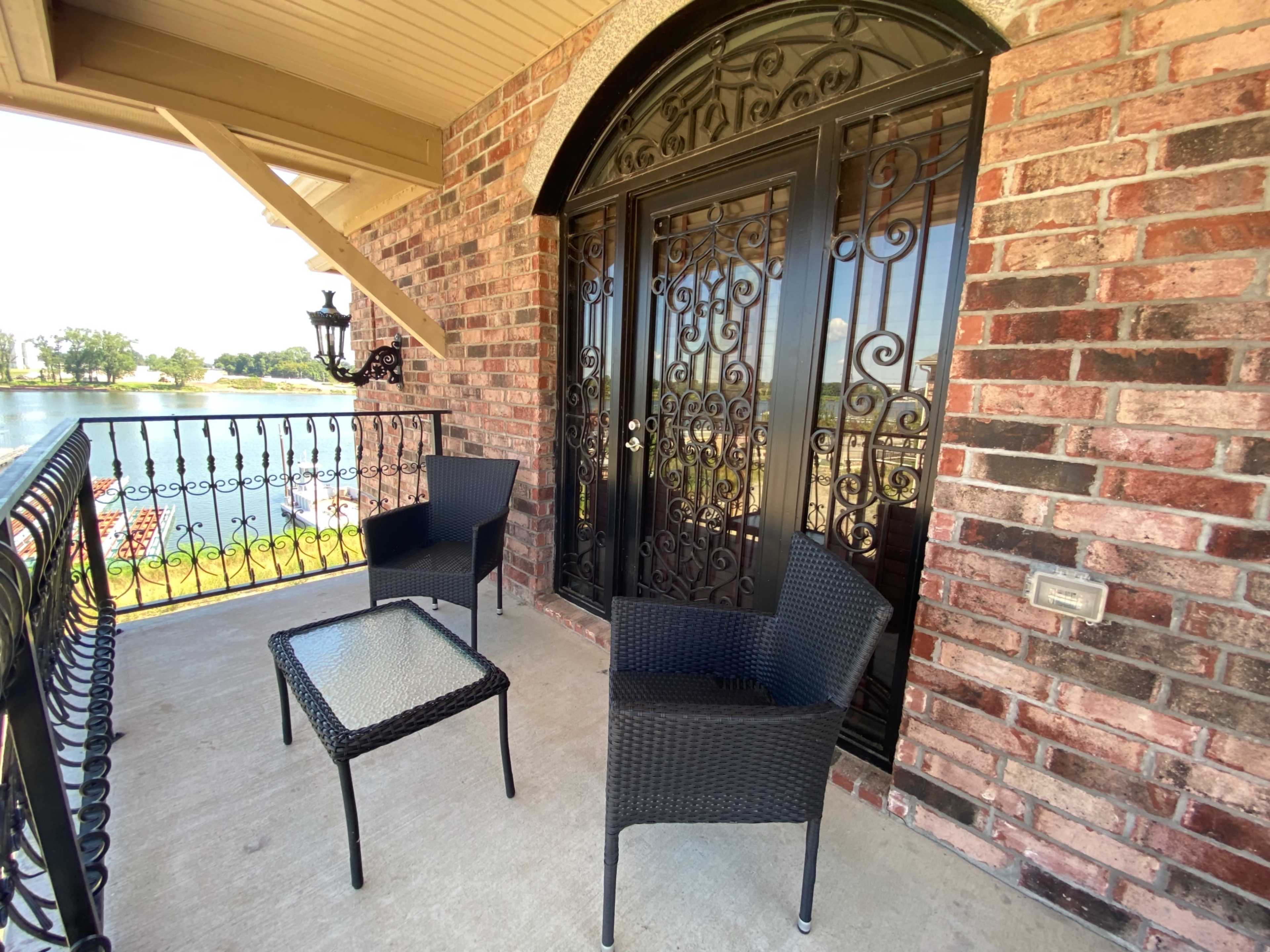 A balcony with two wicker chairs and a glass-topped table, overlooking a body of water.