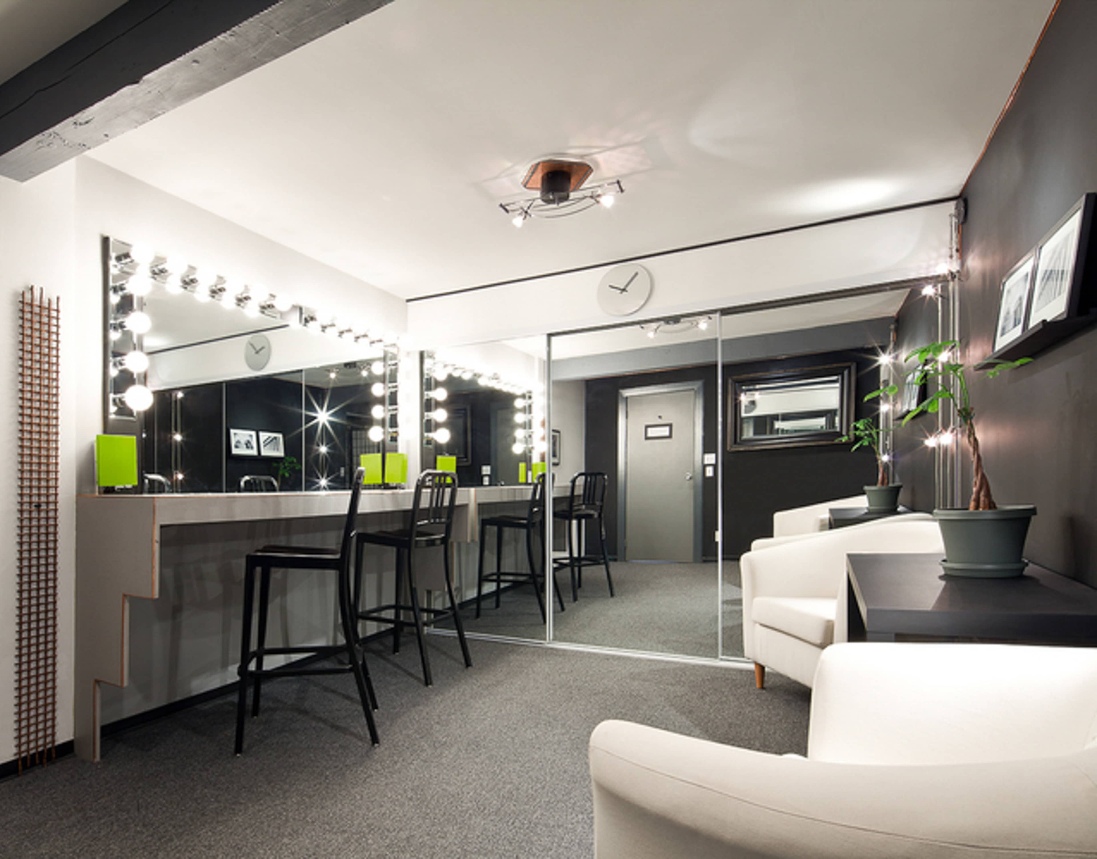 A modern dressing room featuring a row of illuminated mirrors, two bar stools, and a minimalist seating area with a potted plant.