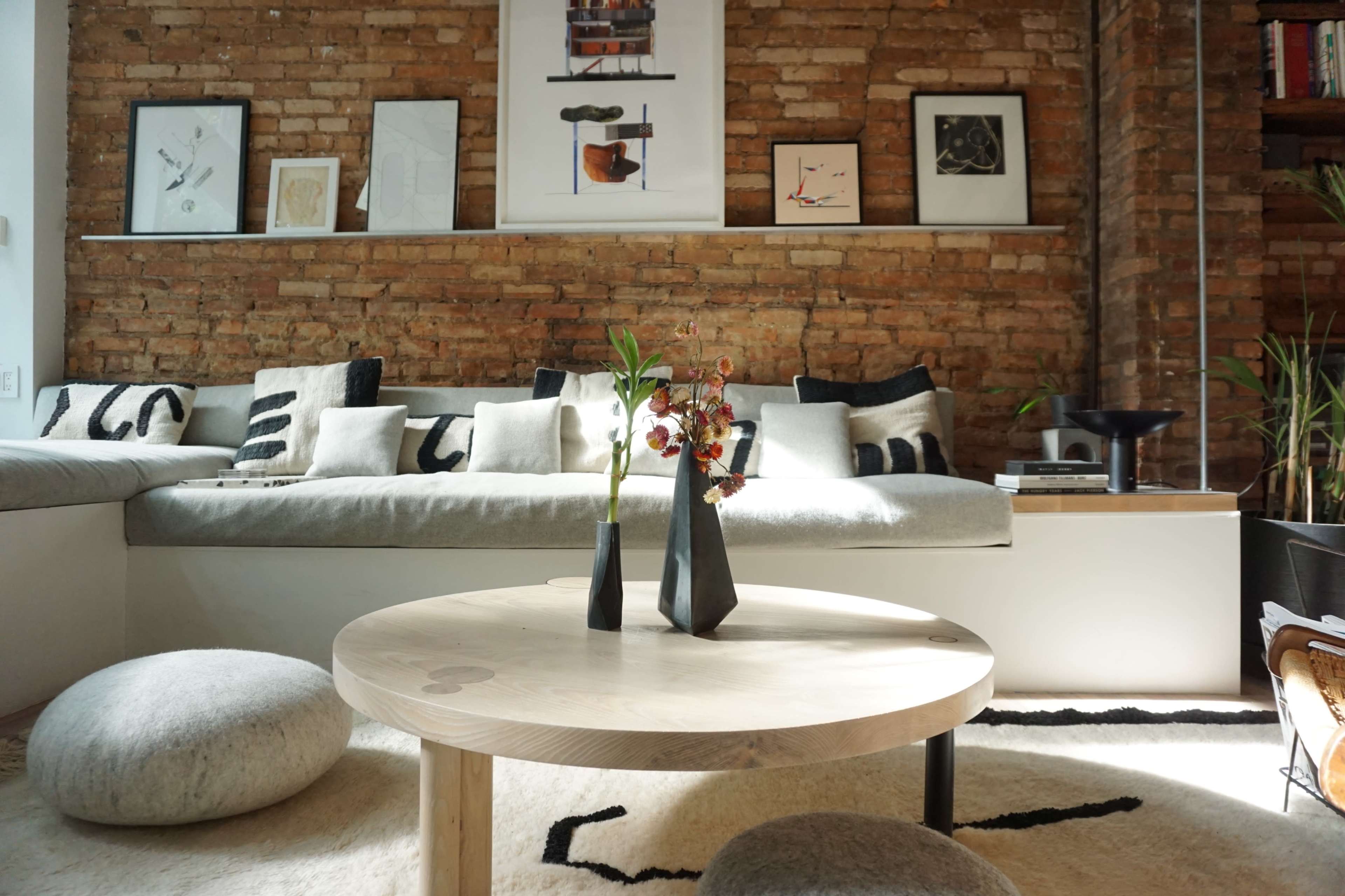 A cozy living room with a brick wall, a large gray sofa adorned with patterned cushions, a round wooden coffee table, and decorative plants.