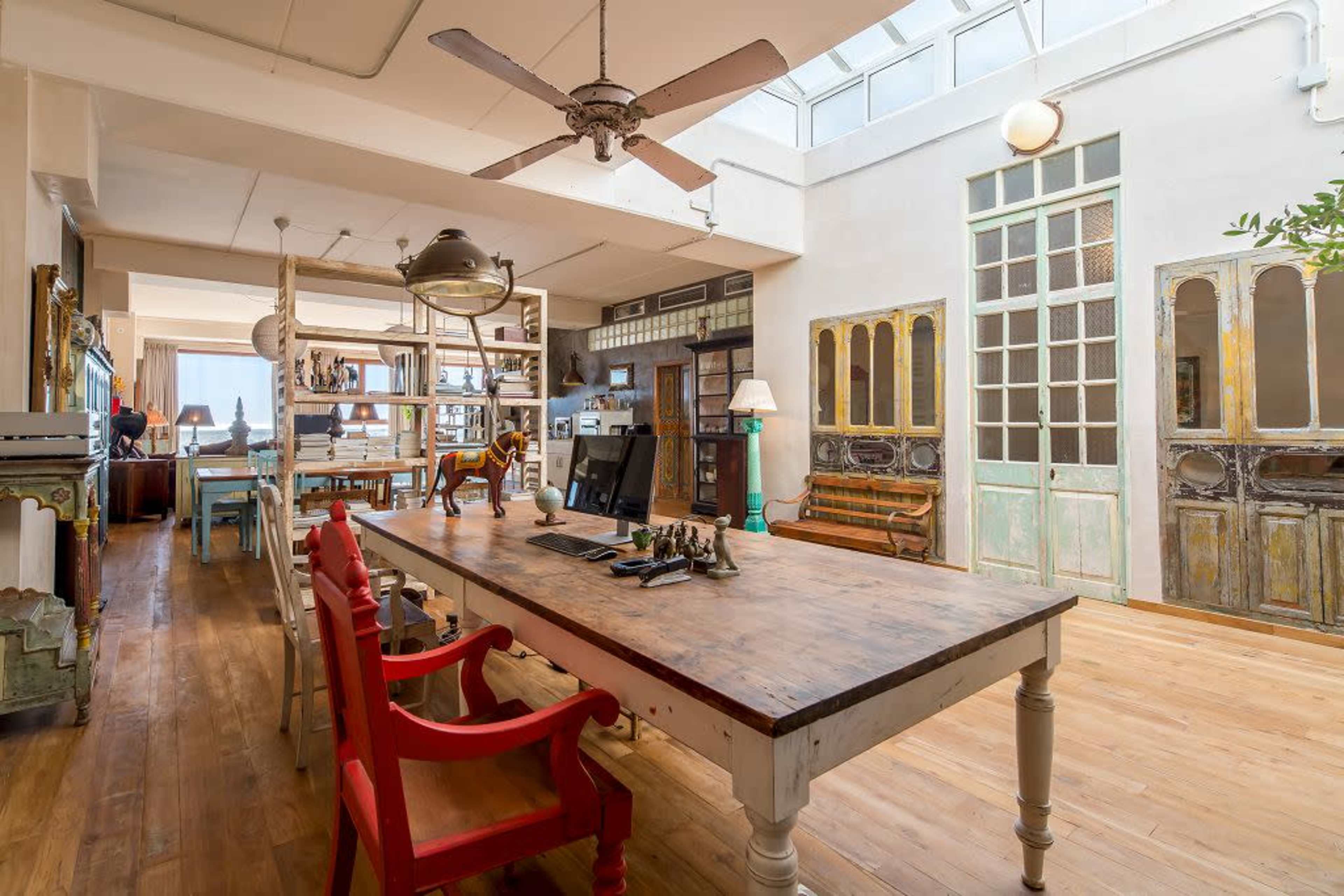 The image depicts a spacious, well-lit interior featuring a wooden table with a computer, colorful chairs, and vintage doors along the walls.