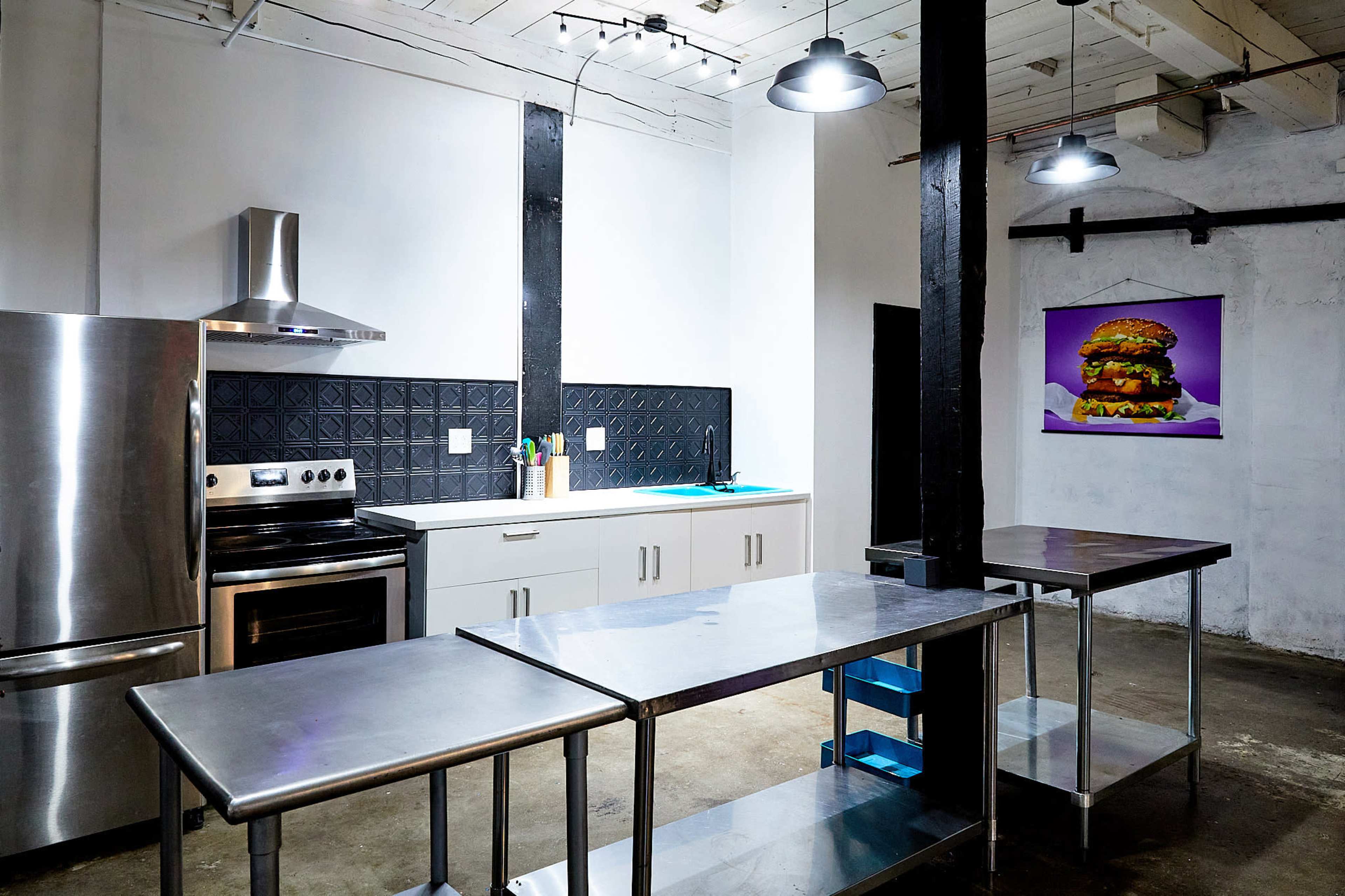 The image shows a modern kitchen with stainless steel appliances, including a stove and refrigerator, along with stainless steel worktables and a large burger poster on the wall.
