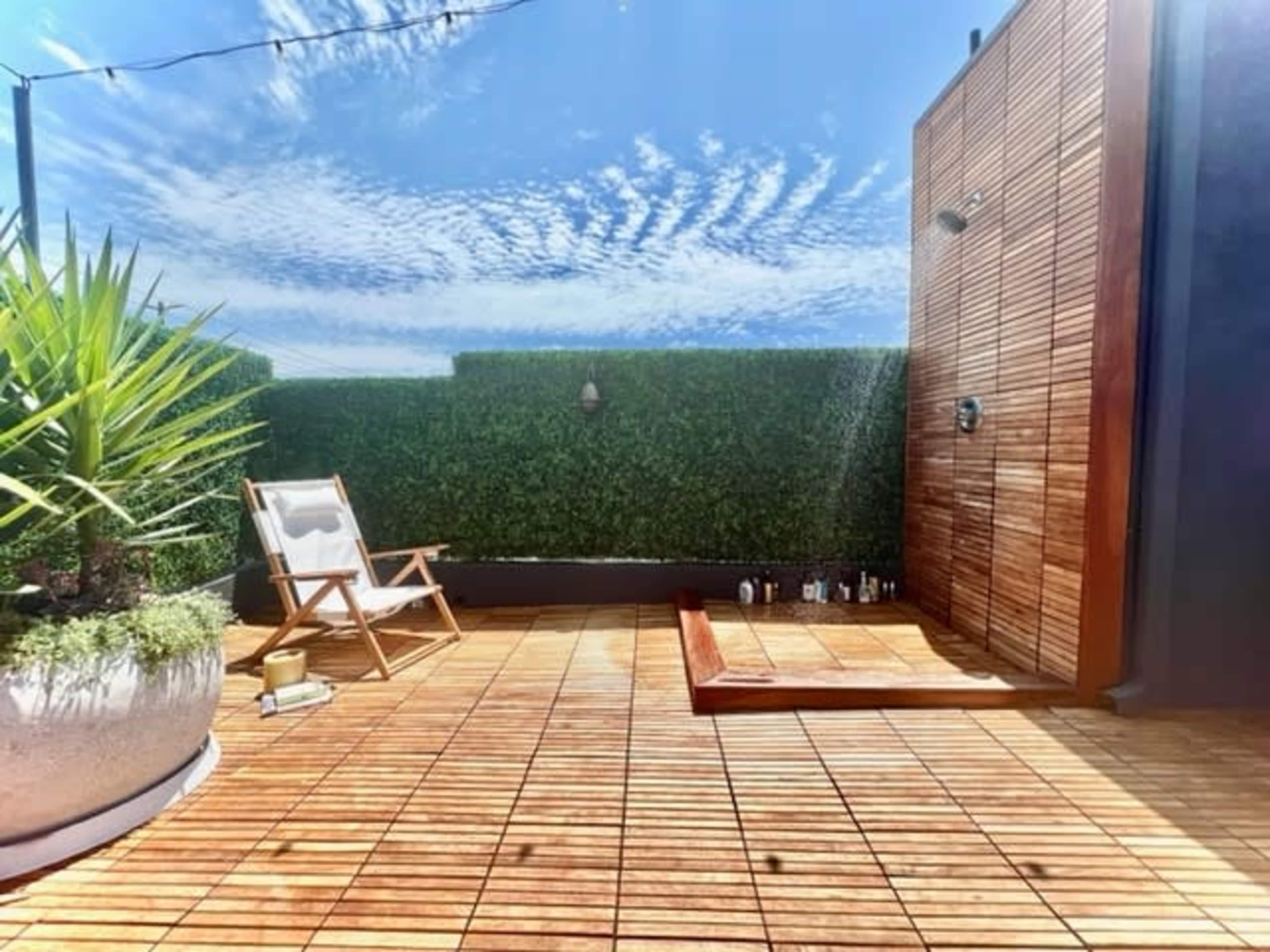A wooden deck with a shower stands on a rooftop terrace surrounded by green hedges under a blue sky with scattered clouds.