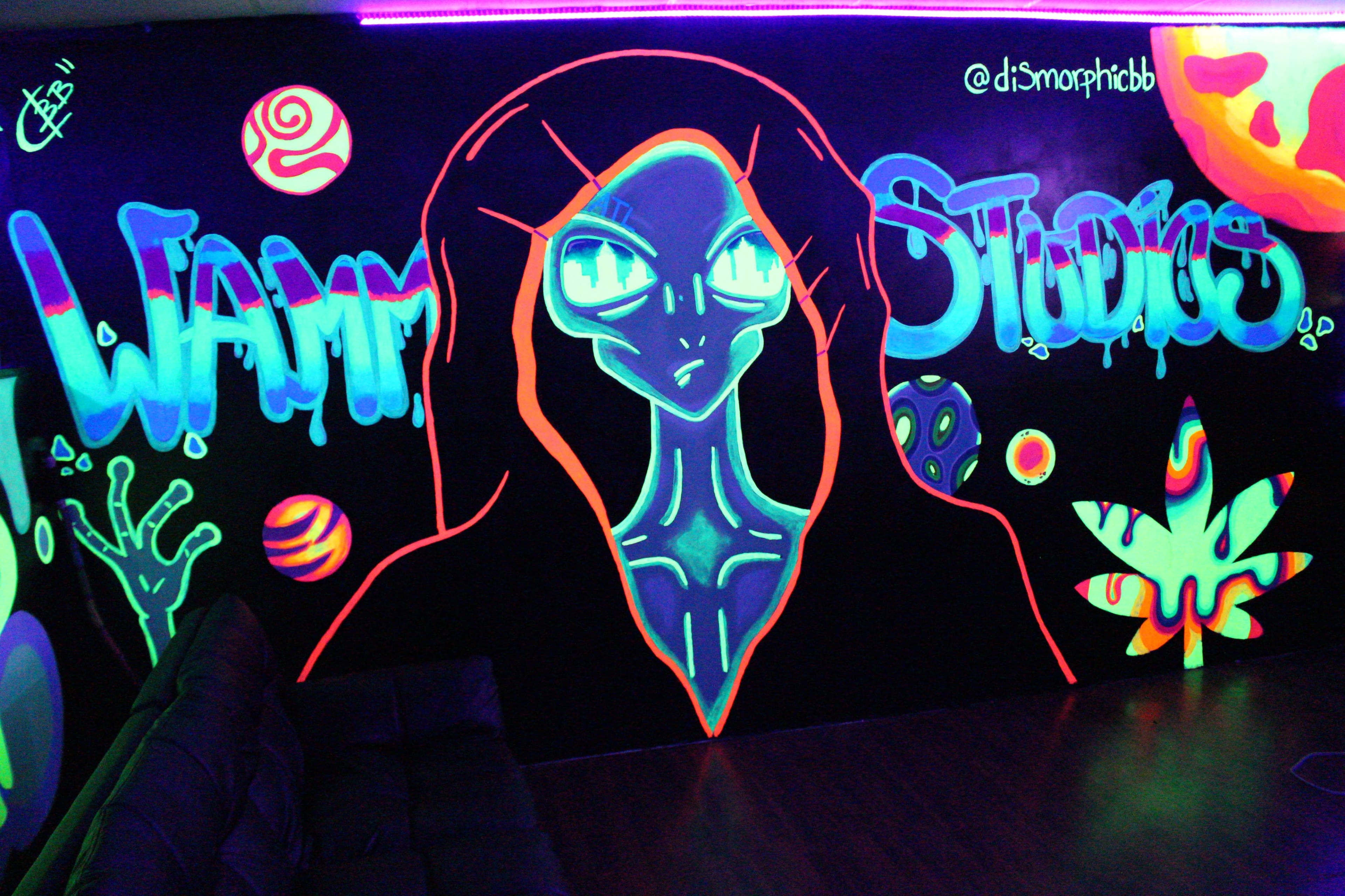 A glowing mural depicting a humanoid figure with large eyes and abstract designs, including planets and a leaf, adorns a dark wall in a studio space.