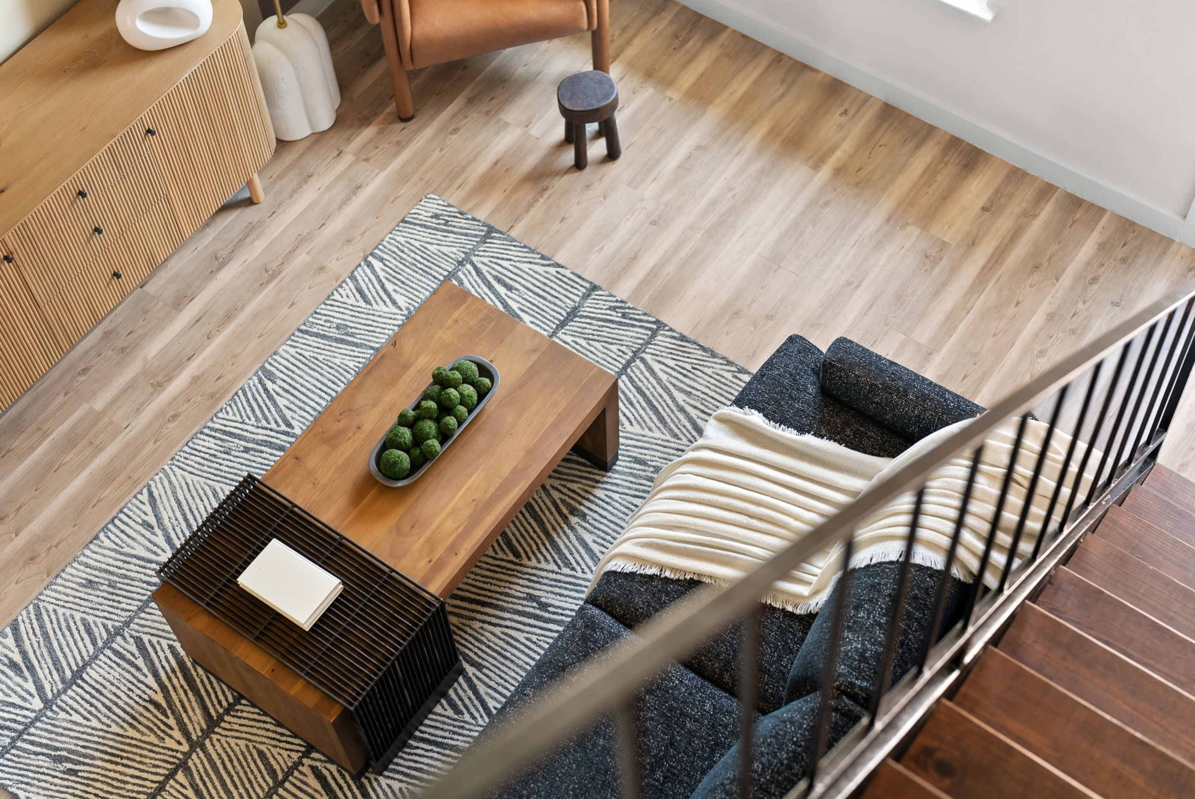 The image shows a modern living room with a wooden coffee table, a dark sofa, a patterned rug, and a staircase in the background.