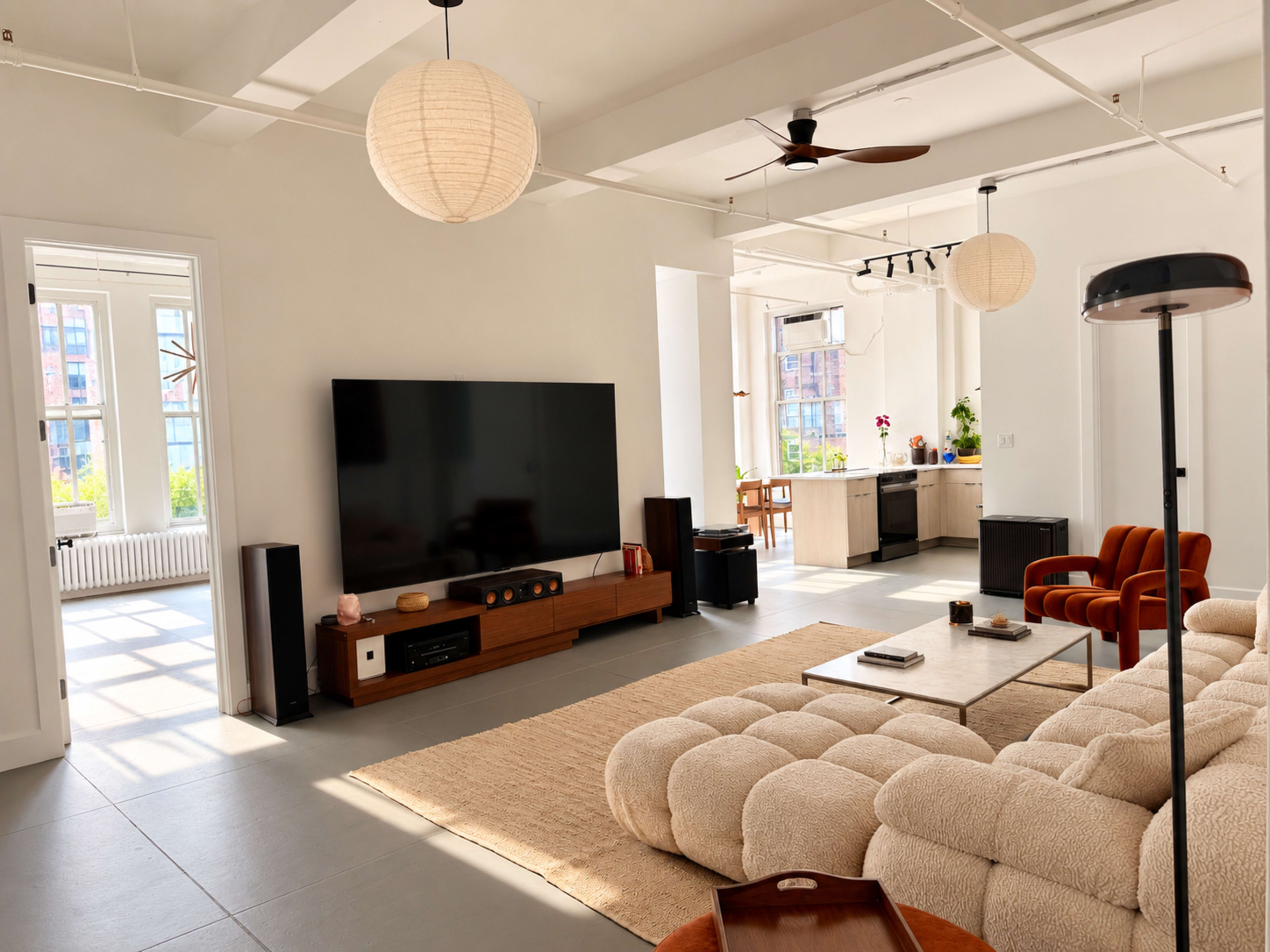 The image shows a modern living room featuring a large television on a wooden stand, a plush sofa, and a kitchen area visible in the background.