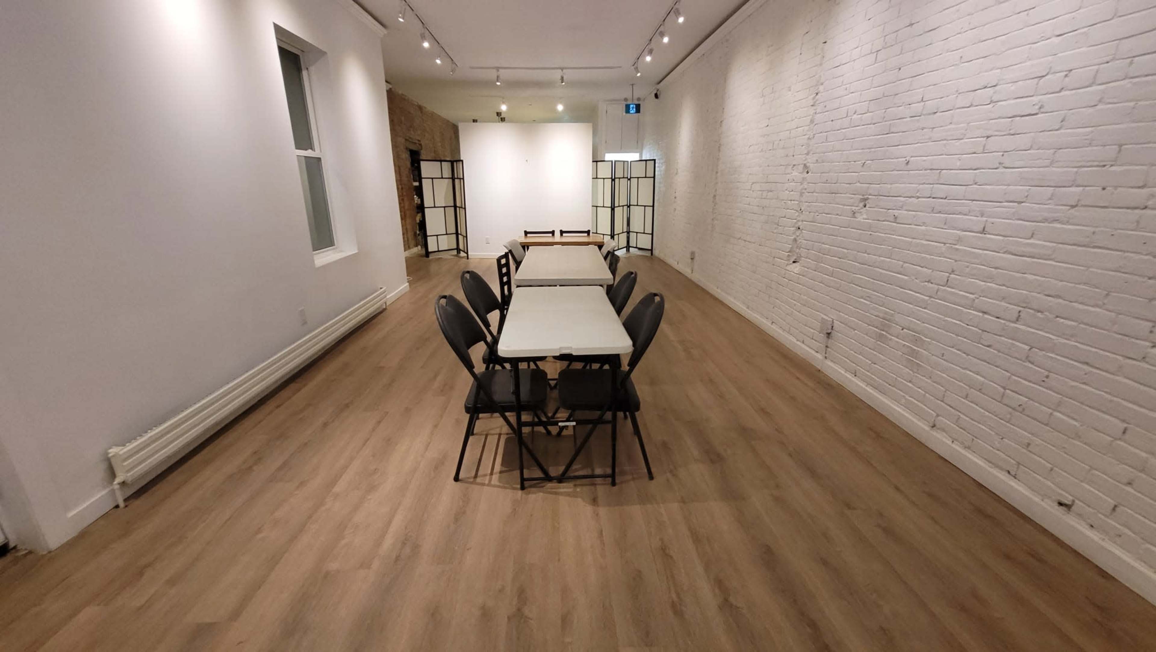 A spacious room features a long row of folding tables surrounded by chairs, with white brick walls and wooden flooring.