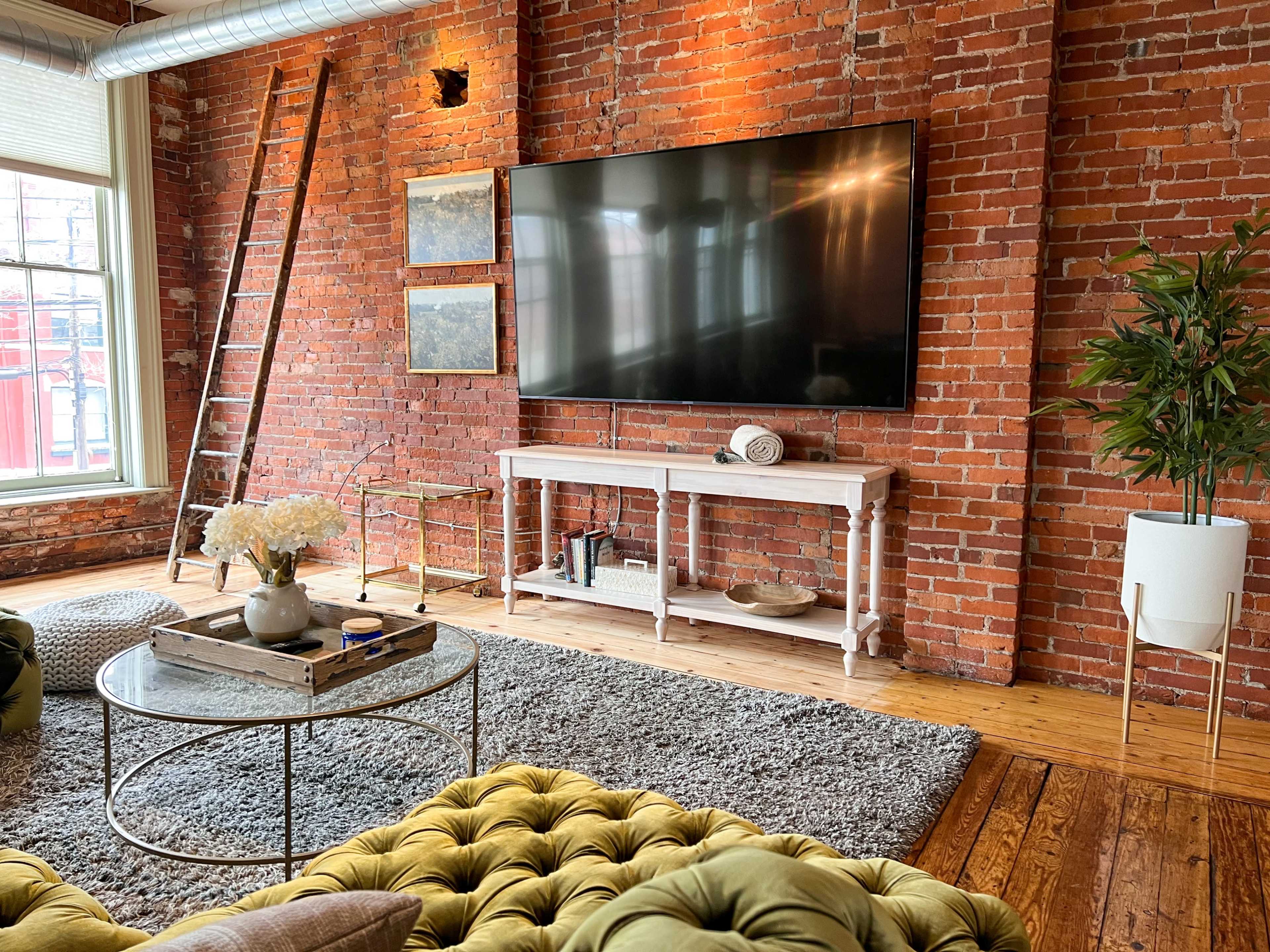 The scene features a living room with exposed brick walls, a large television mounted on the wall, and a decorative coffee table amidst cozy seating arrangements.