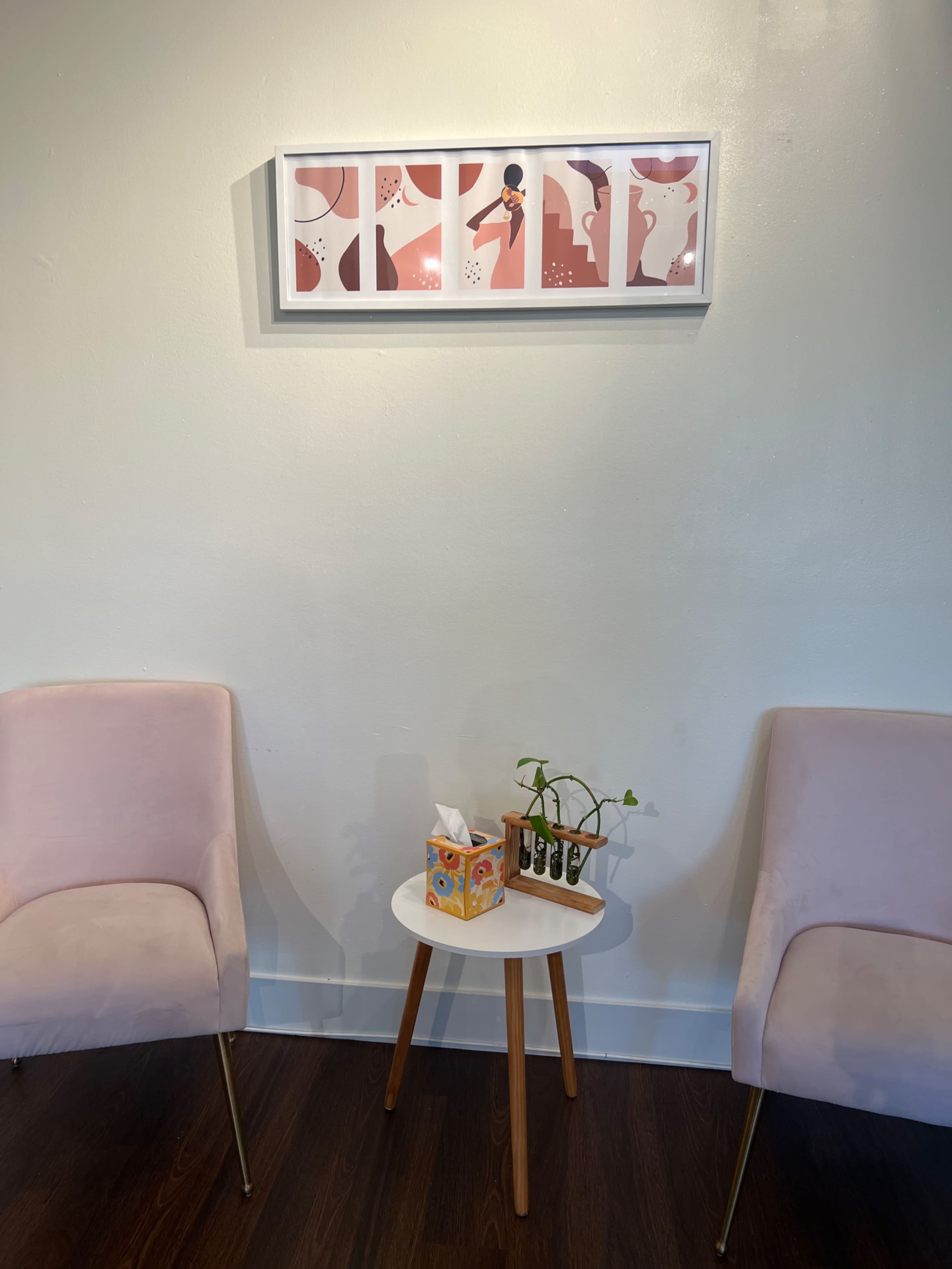 The image shows two pink chairs flanking a small white table with a tissue box and a potted plant, beneath a framed artwork featuring abstract figures on the wall.