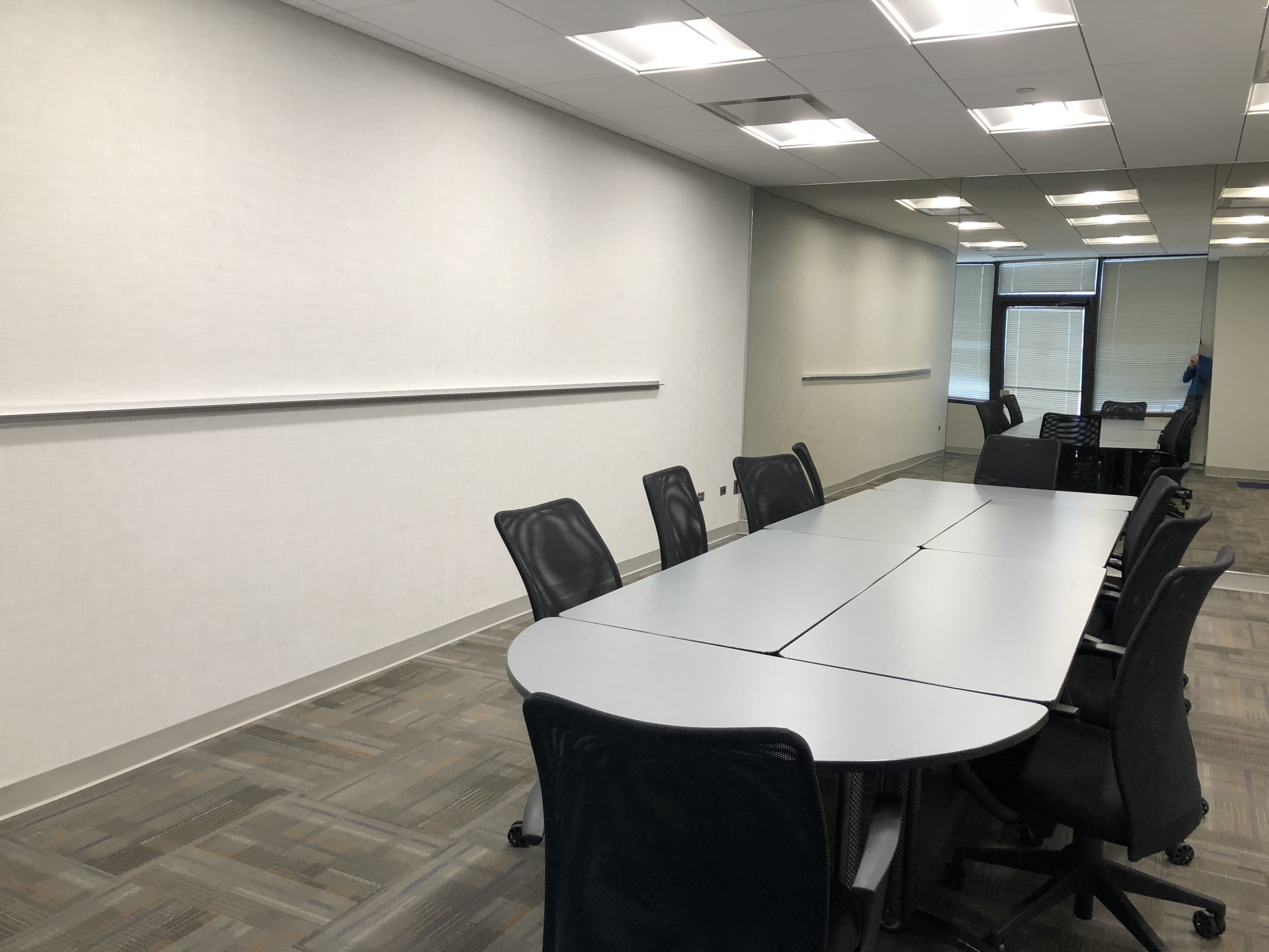 A large, rectangular conference table with black mesh chairs is positioned in a brightly lit meeting room with plain white walls.