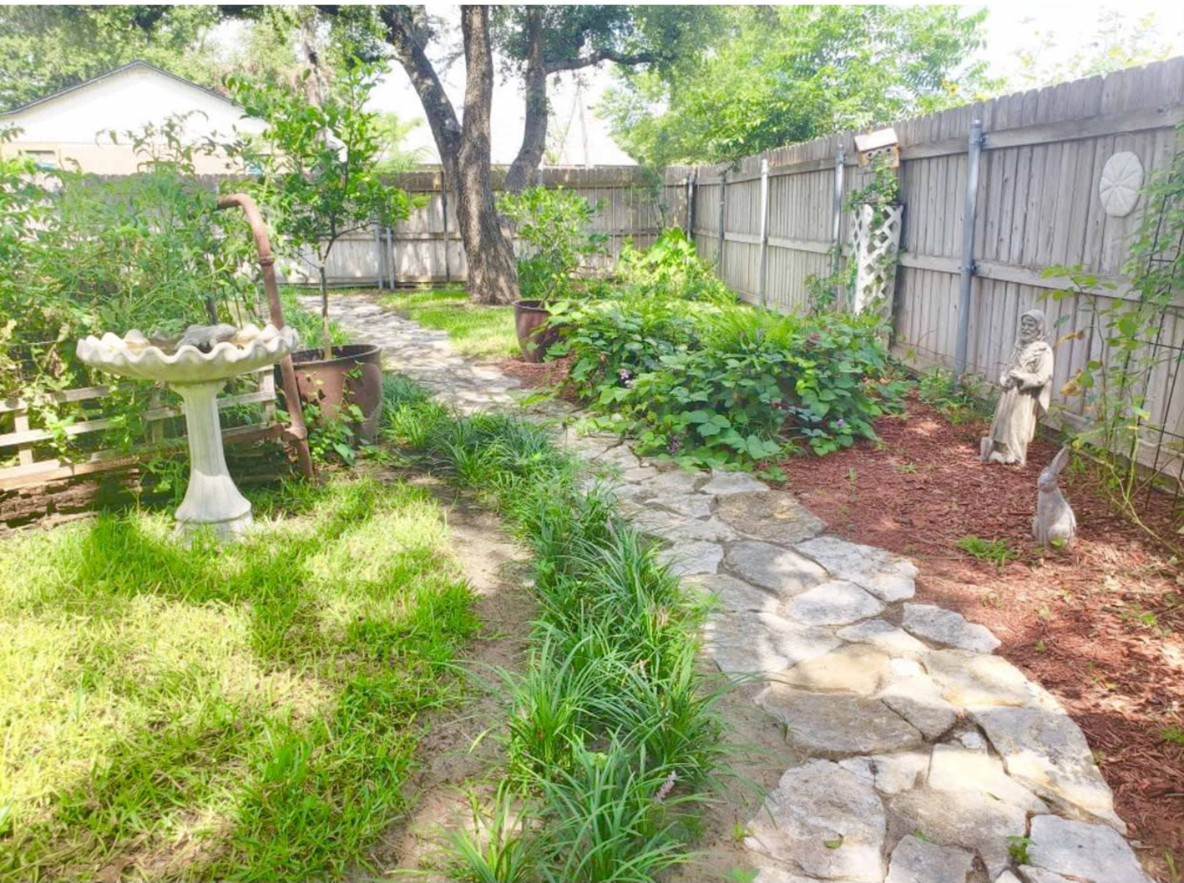 A stone pathway winds through a garden featuring greenery, potted plants, a birdbath, and a statue.