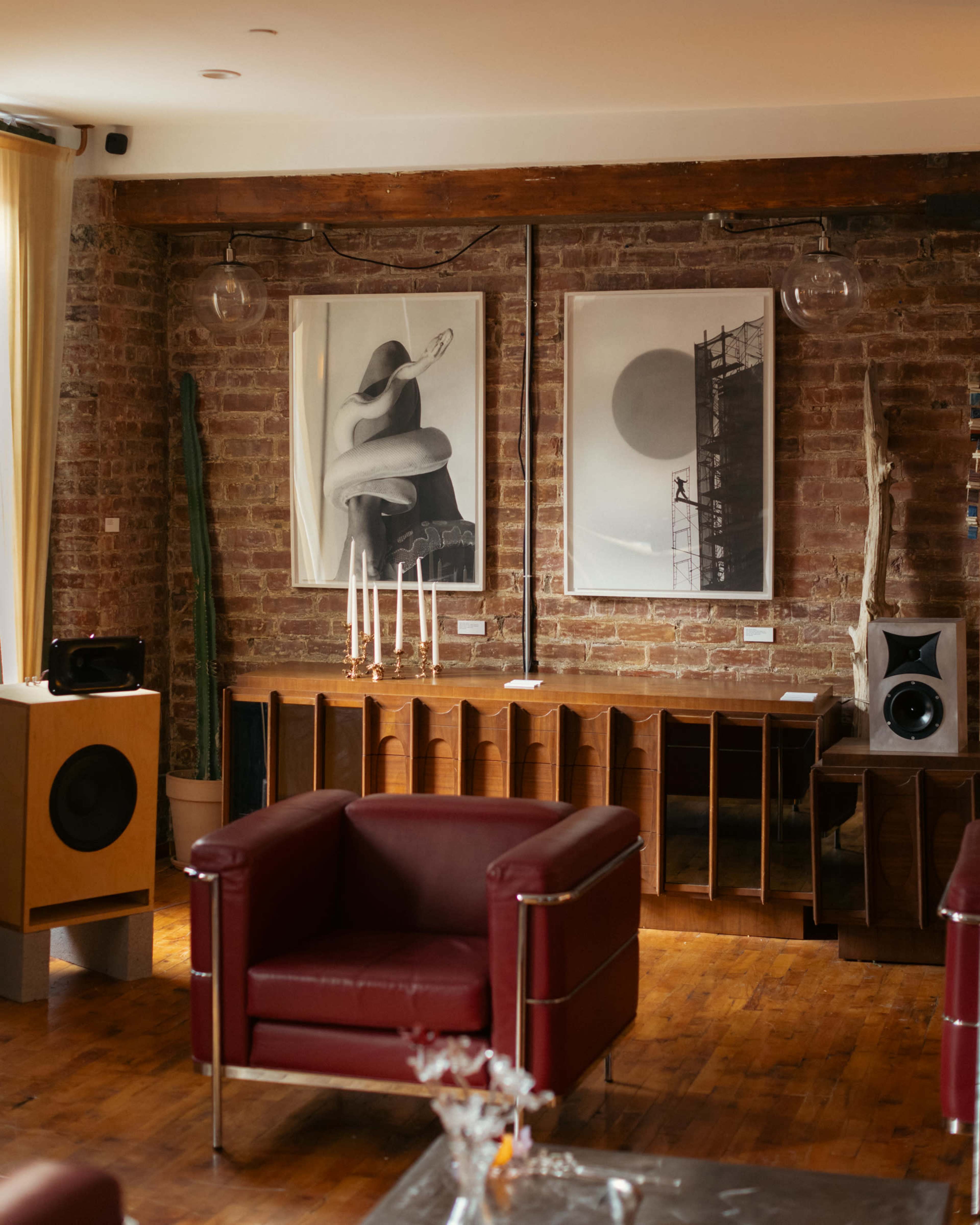 The image shows a stylish interior featuring two large framed artworks on a brick wall, a red armchair, wooden furniture, and a decorative plant.