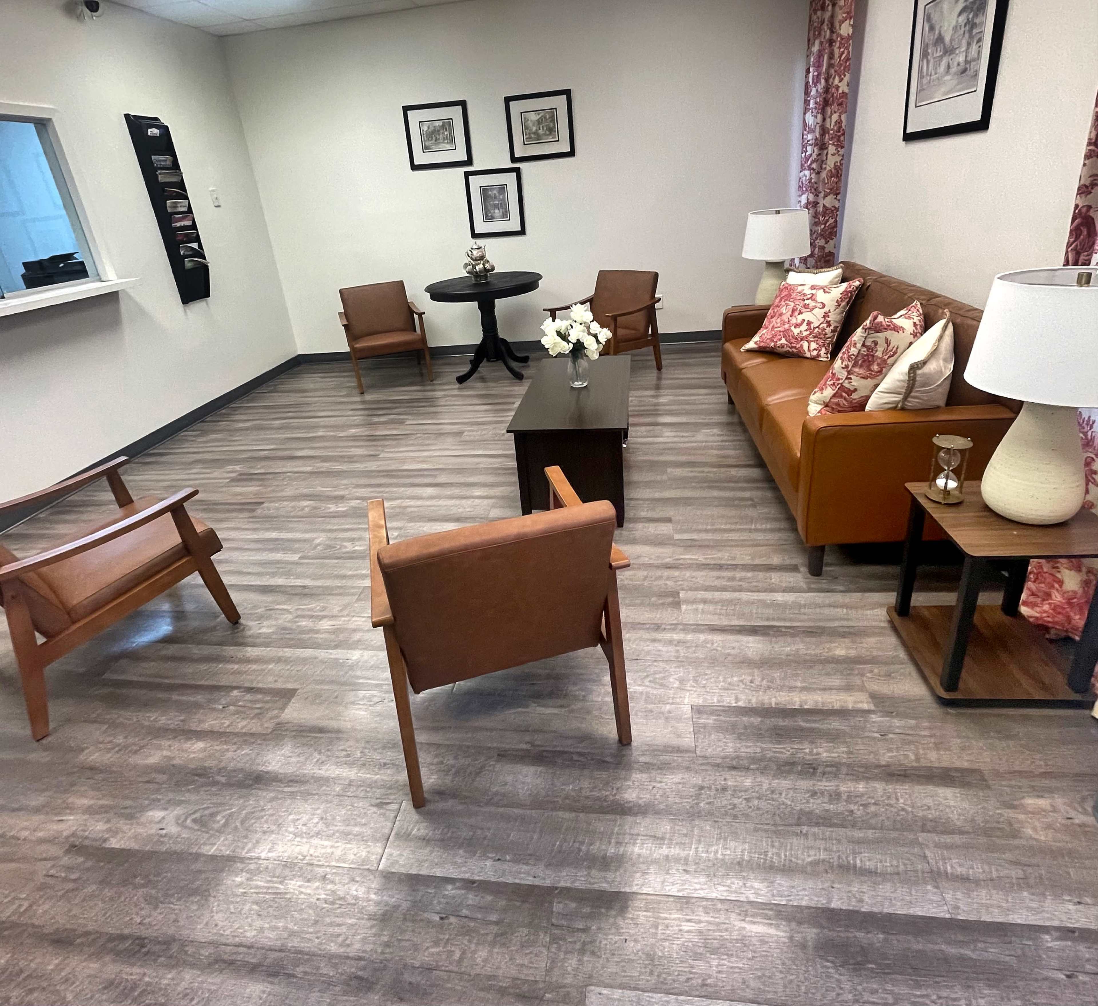The image shows a small waiting room with a brown leather sofa, two wooden chairs, a round table, and decorative photos on the walls.