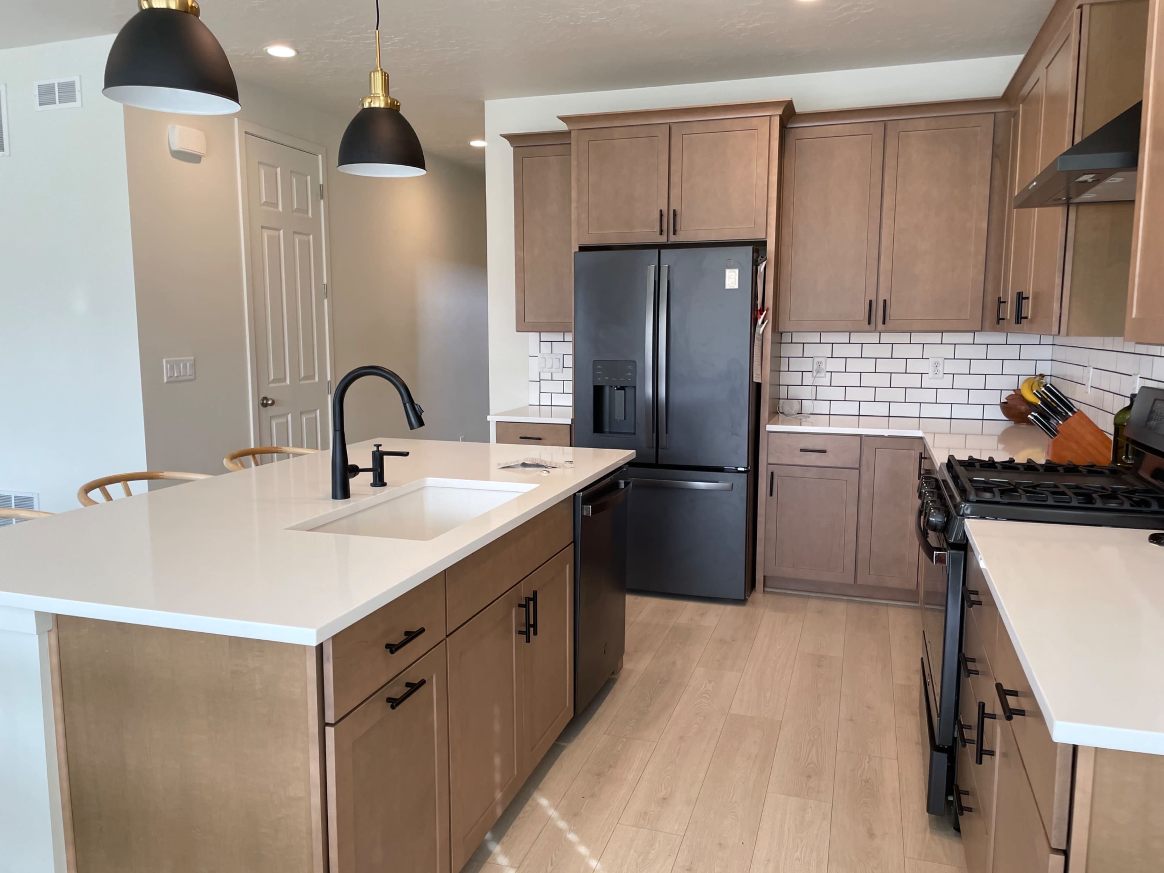 A modern kitchen featuring wooden cabinets, a black refrigerator, a white countertop, and a sink island with black pendant lights overhead.