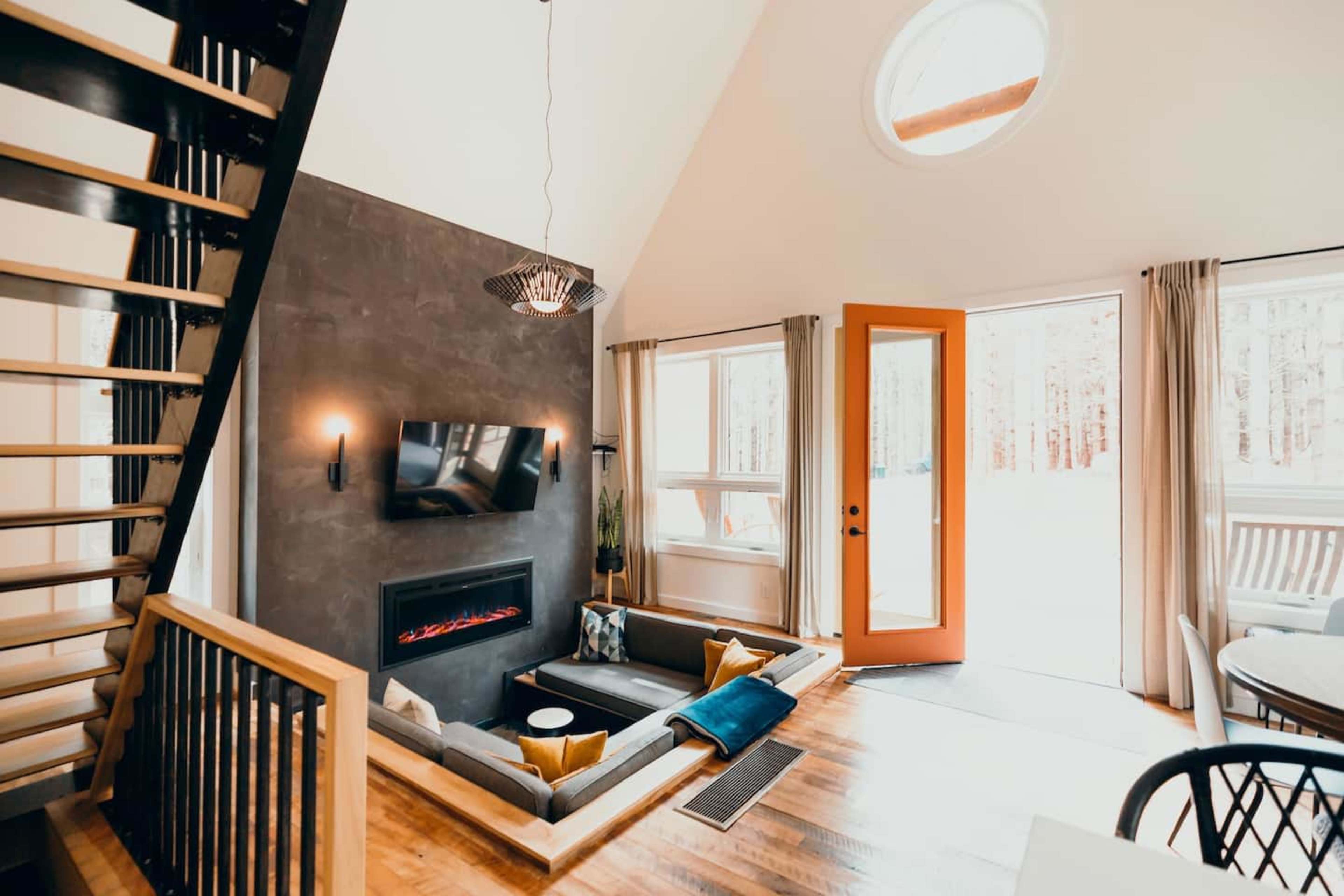 A modern living room with a fireplace, a large wall-mounted TV, and a view of a patio area through orange double doors.