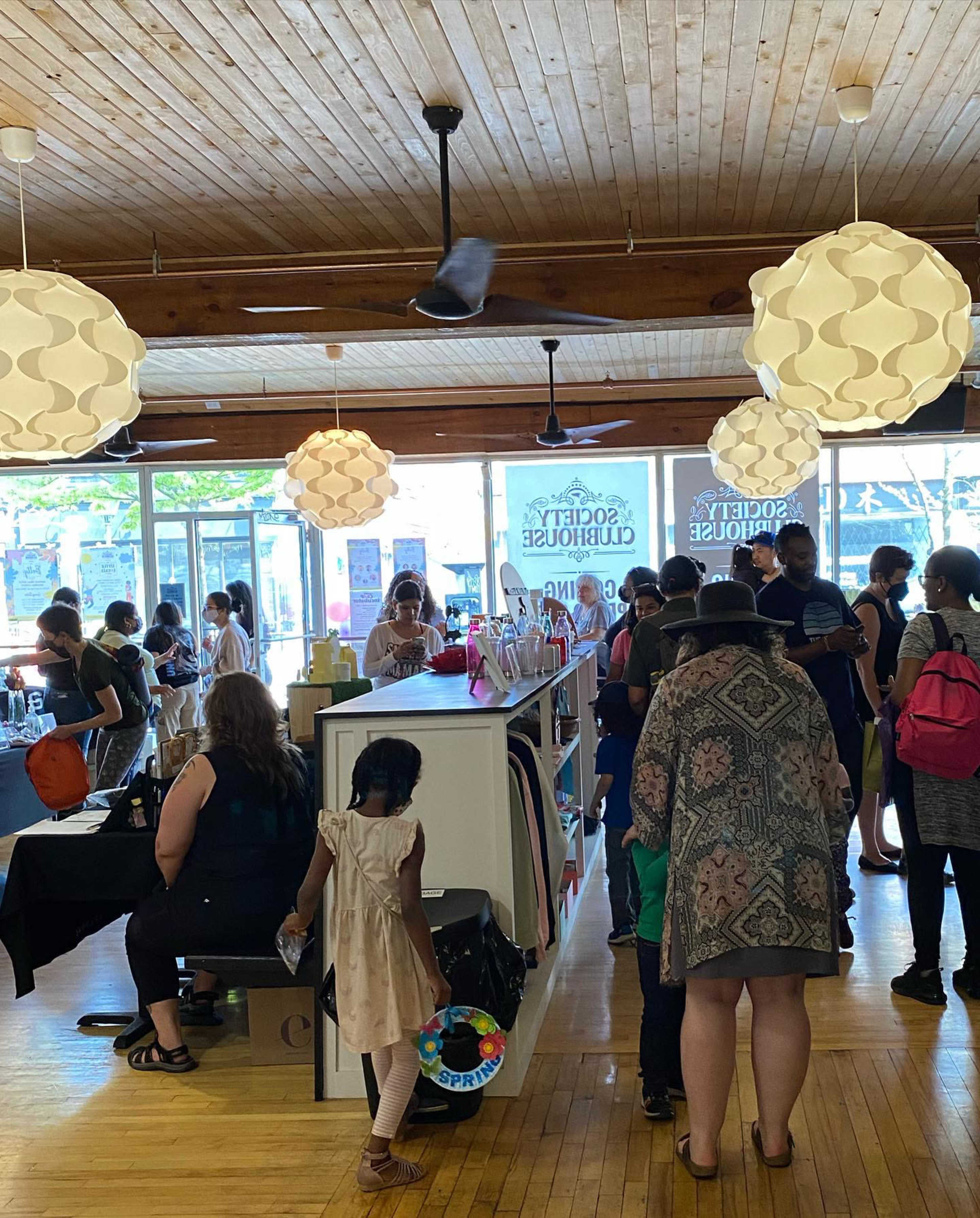 A busy retail space filled with customers browsing products, with large windows and hanging light fixtures overhead.