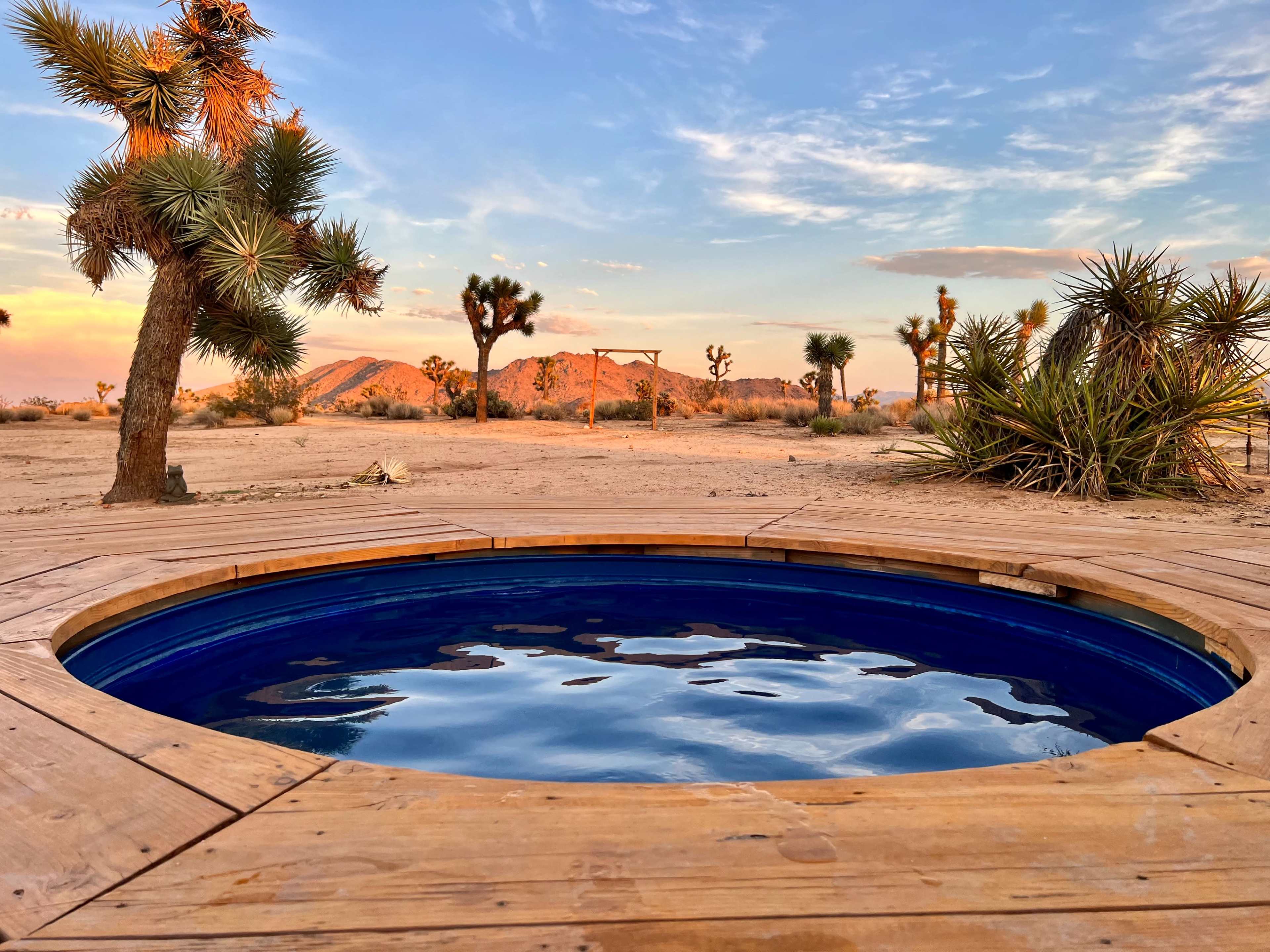 A circular hot tub is set on a wooden deck in a desert landscape with Joshua trees and mountains in the background during sunset.