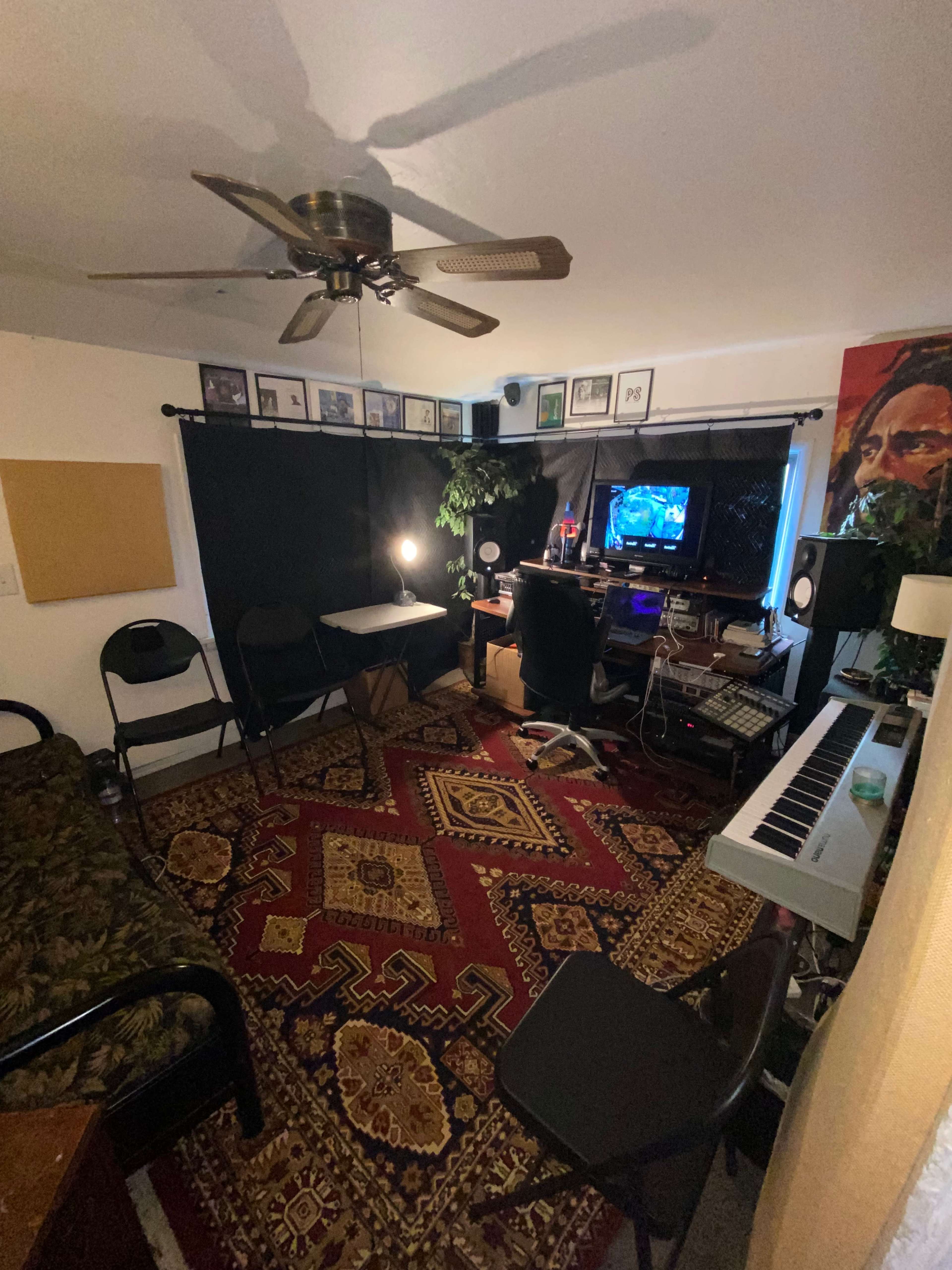 The image shows a small room arranged as a workspace with a computer desk, musical keyboard, and a patterned rug, along with a ceiling fan and framed pictures on the walls.