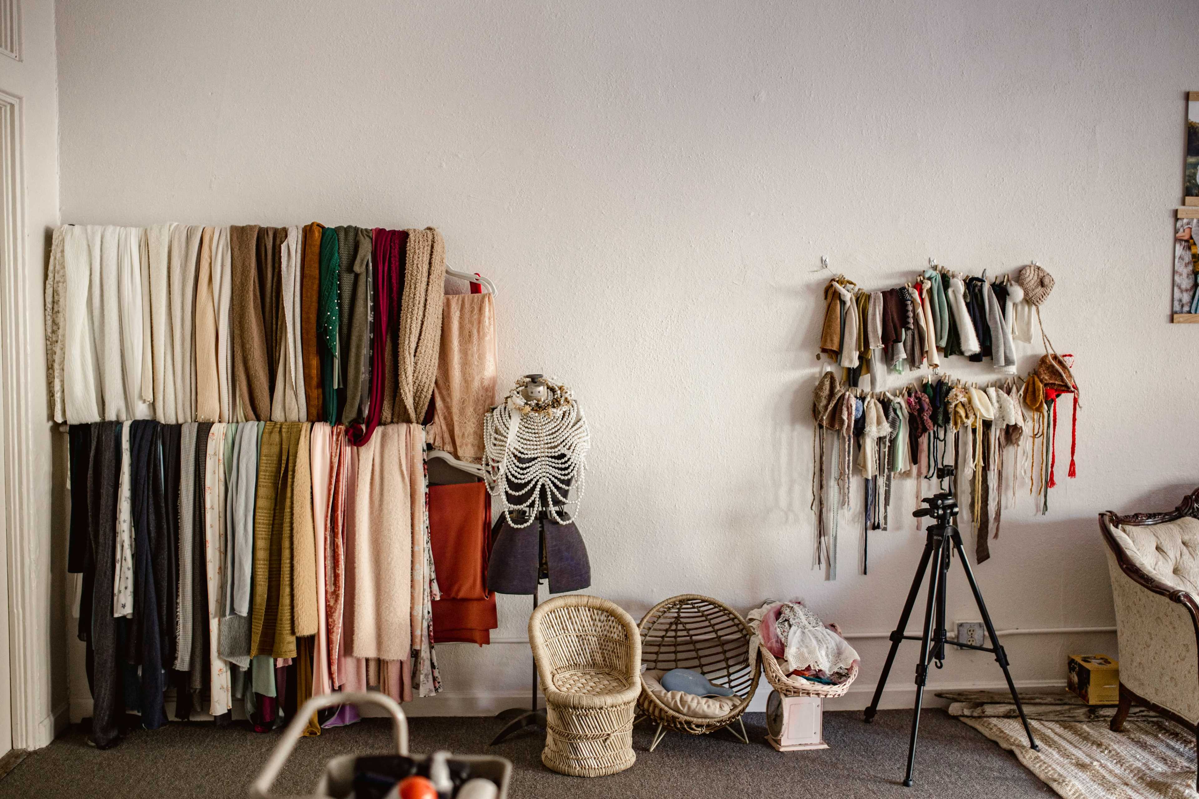 The image shows a room with neatly arranged textile fabrics hanging on the left wall and various accessories displayed on the right, along with a wicker basket and a tripod positioned on the floor.