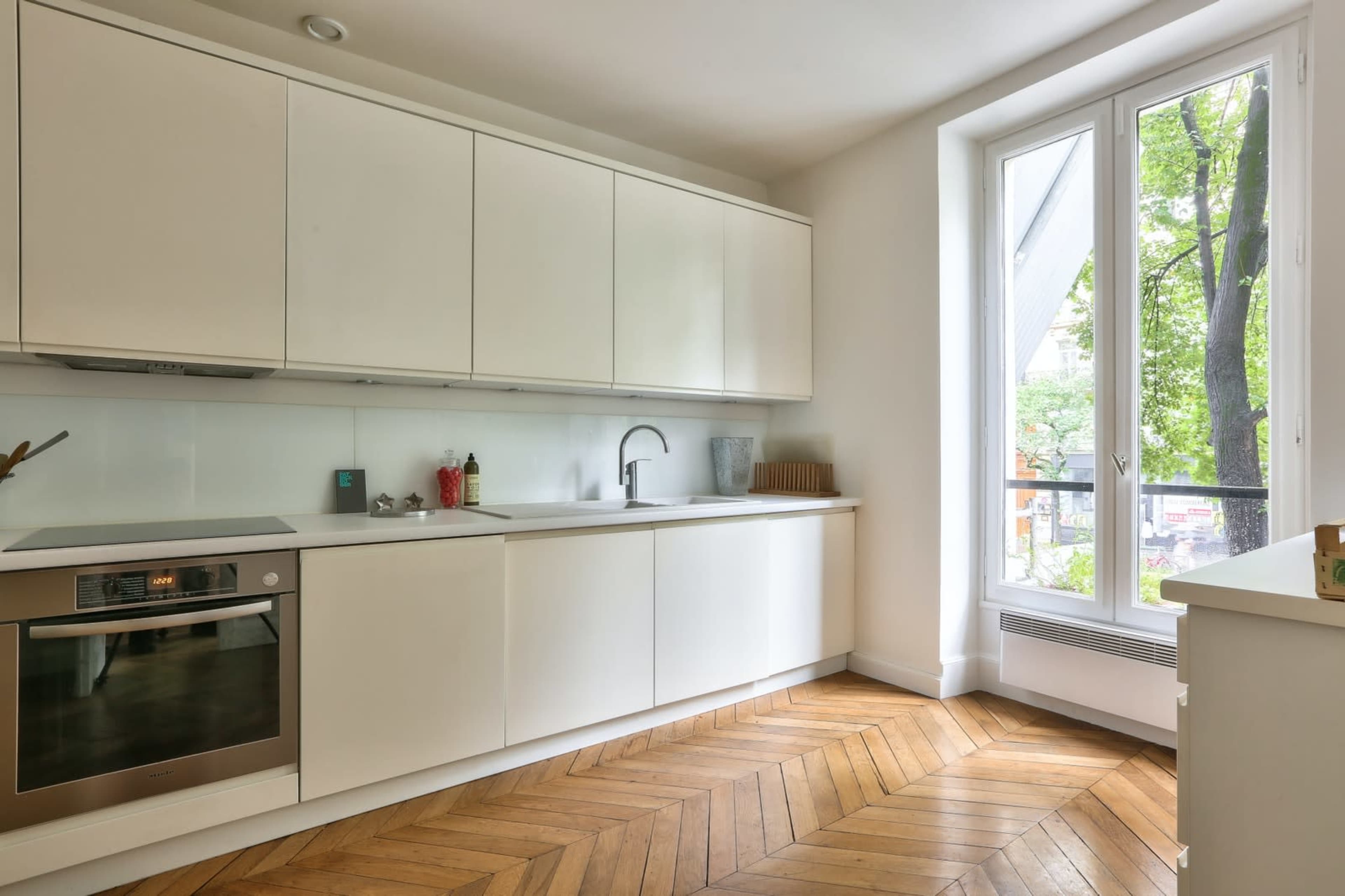 The image shows a modern kitchen with white cabinetry, stainless steel appliances, and a window that overlooks greenery.