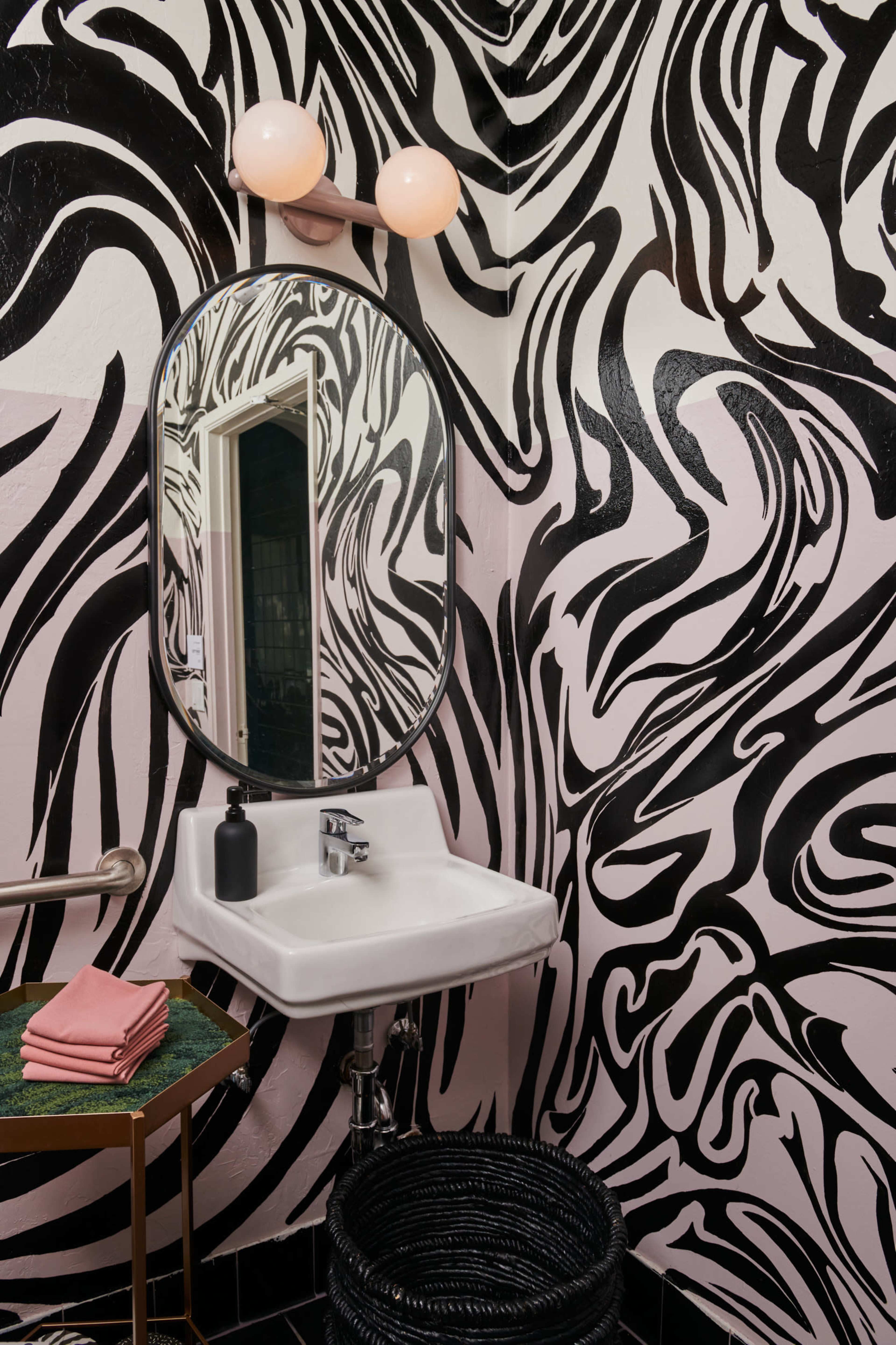 A bathroom with a striking black and pink swirl pattern on the walls, a modern oval mirror above a white sink, and a small green table with folded towels nearby.