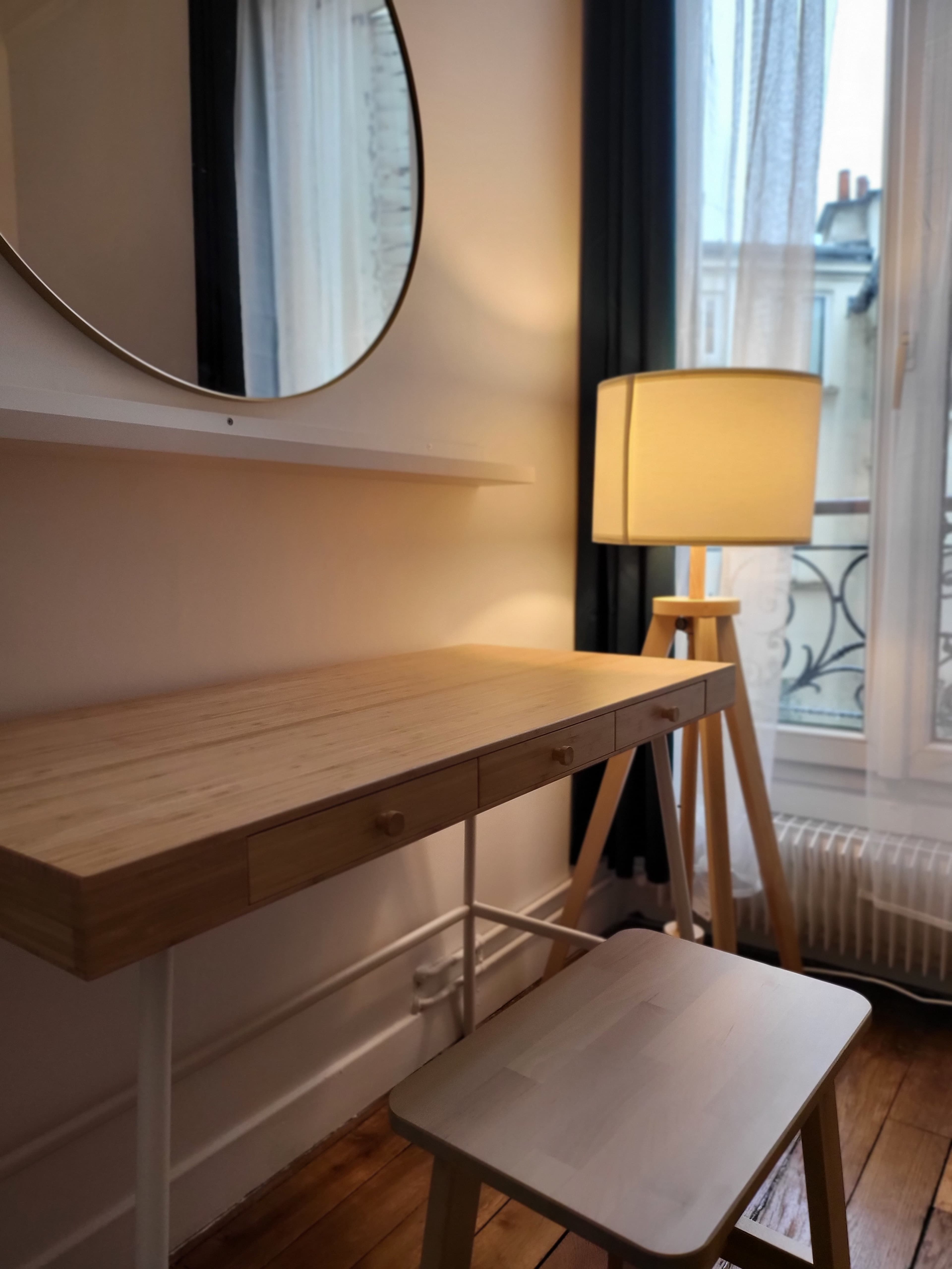 The image shows a minimalist workspace featuring a wooden desk and stool with a round mirror above, next to a tall lamp, all set by a window with sheer curtains.