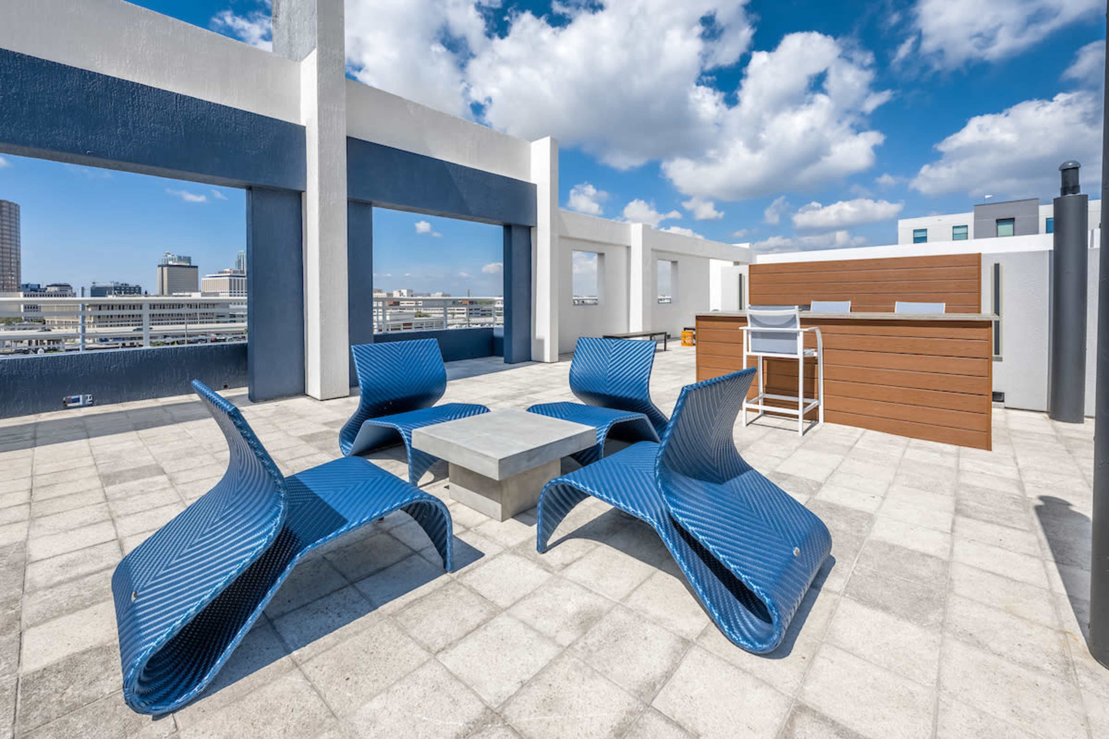 A rooftop terrace featuring modern blue lounge chairs arranged around a concrete table, with a wooden bar area in the background and a clear blue sky above.