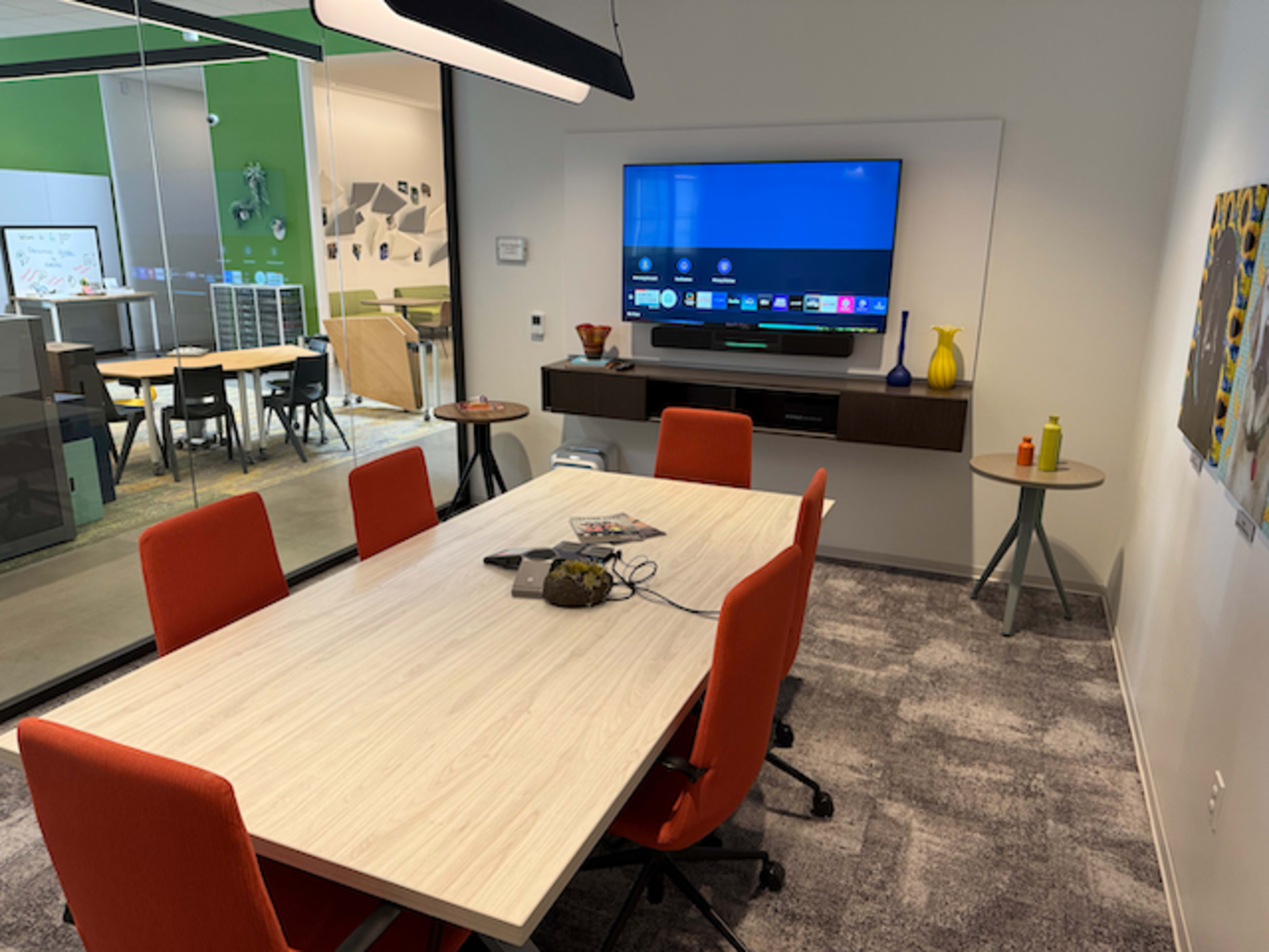 A conference room features a rectangular table surrounded by orange chairs, a wall-mounted TV, and various items on the table.
