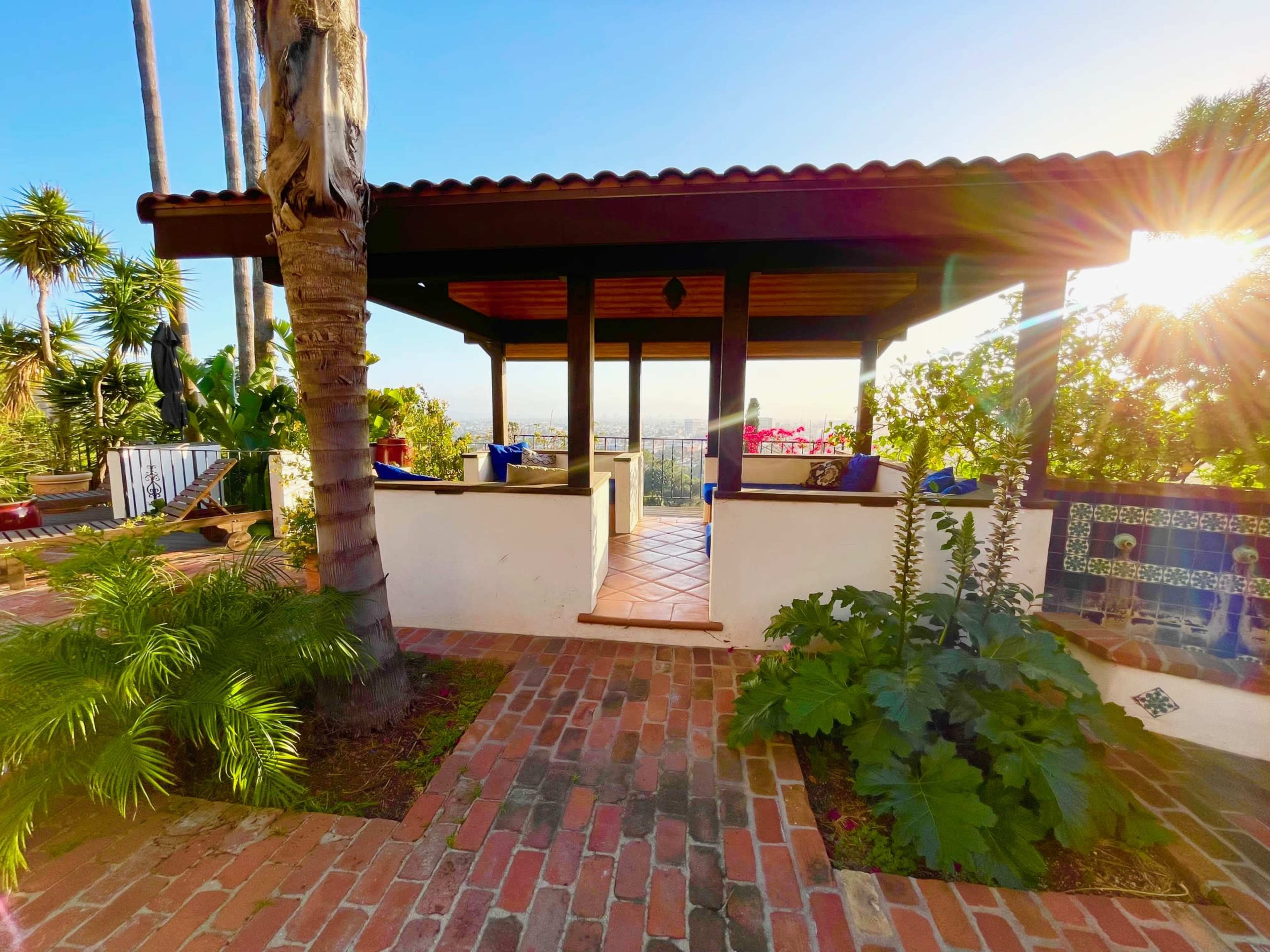 The image shows a patio with a wooden pergola surrounded by tropical plants and a view of a cityscape in the background.