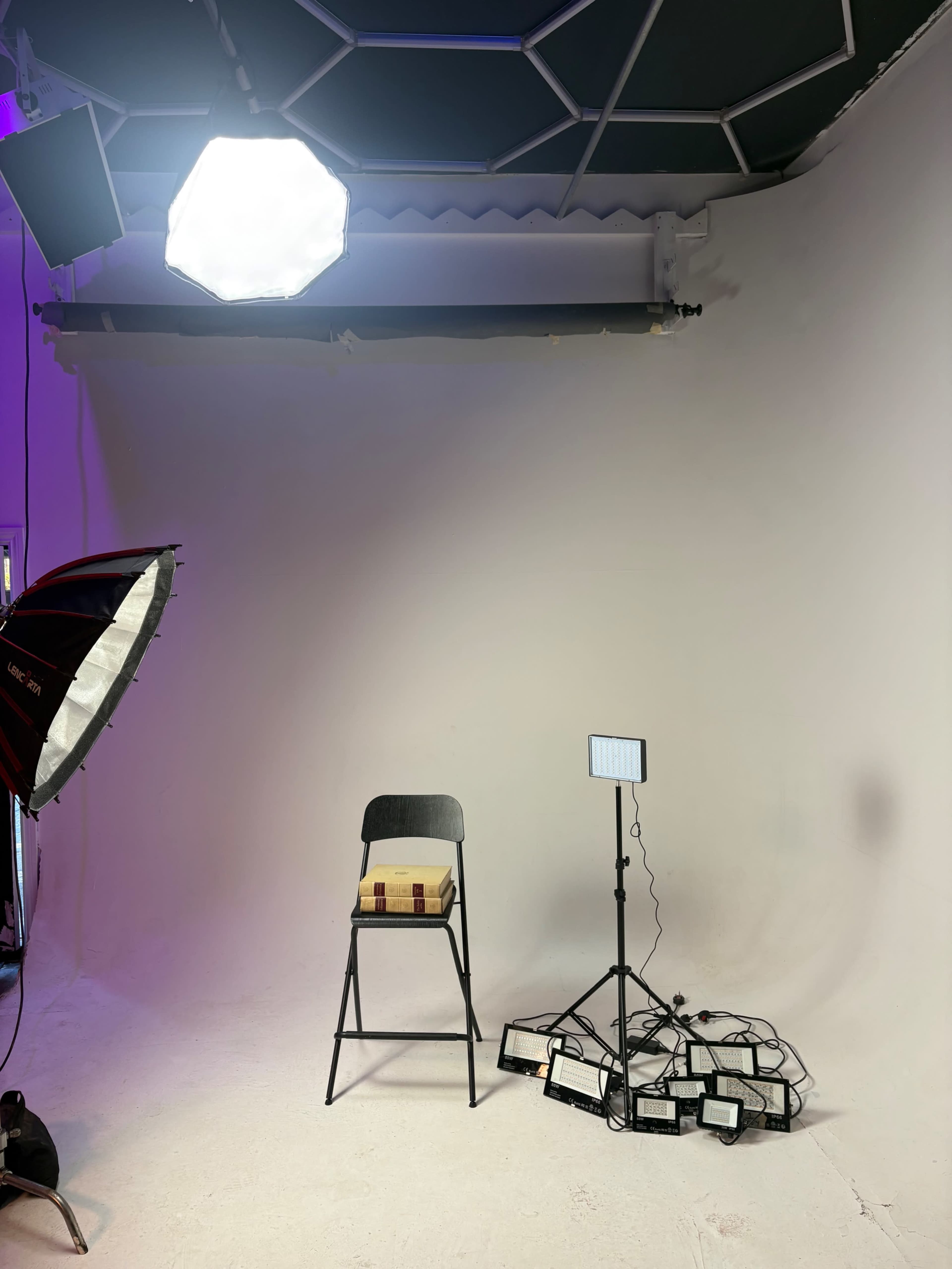 The image shows a photography studio with a chair, a light setup featuring an umbrella softbox and several LED panel lights, against a plain white backdrop.