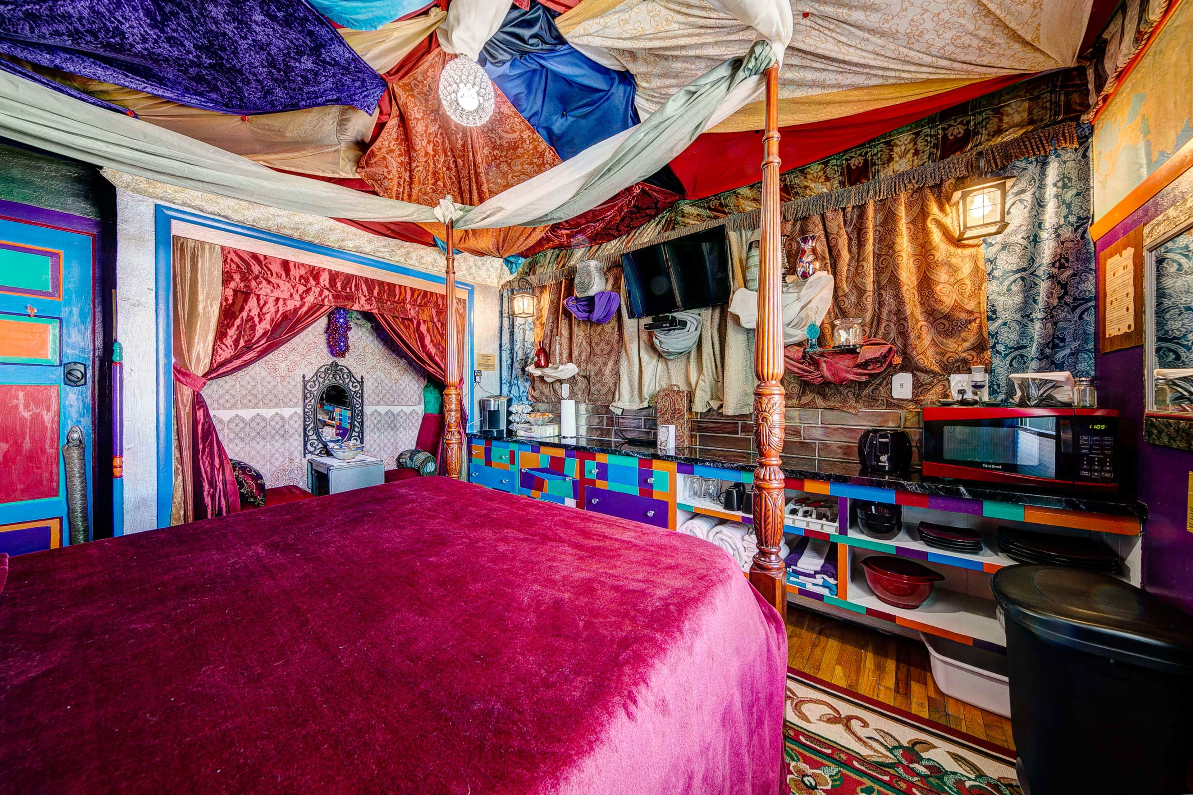 The image shows a vibrant and eclectic bedroom with a four-poster bed draped in colorful fabrics, a mix of decorative wall treatments, and various furnishings including a microwave and a television.