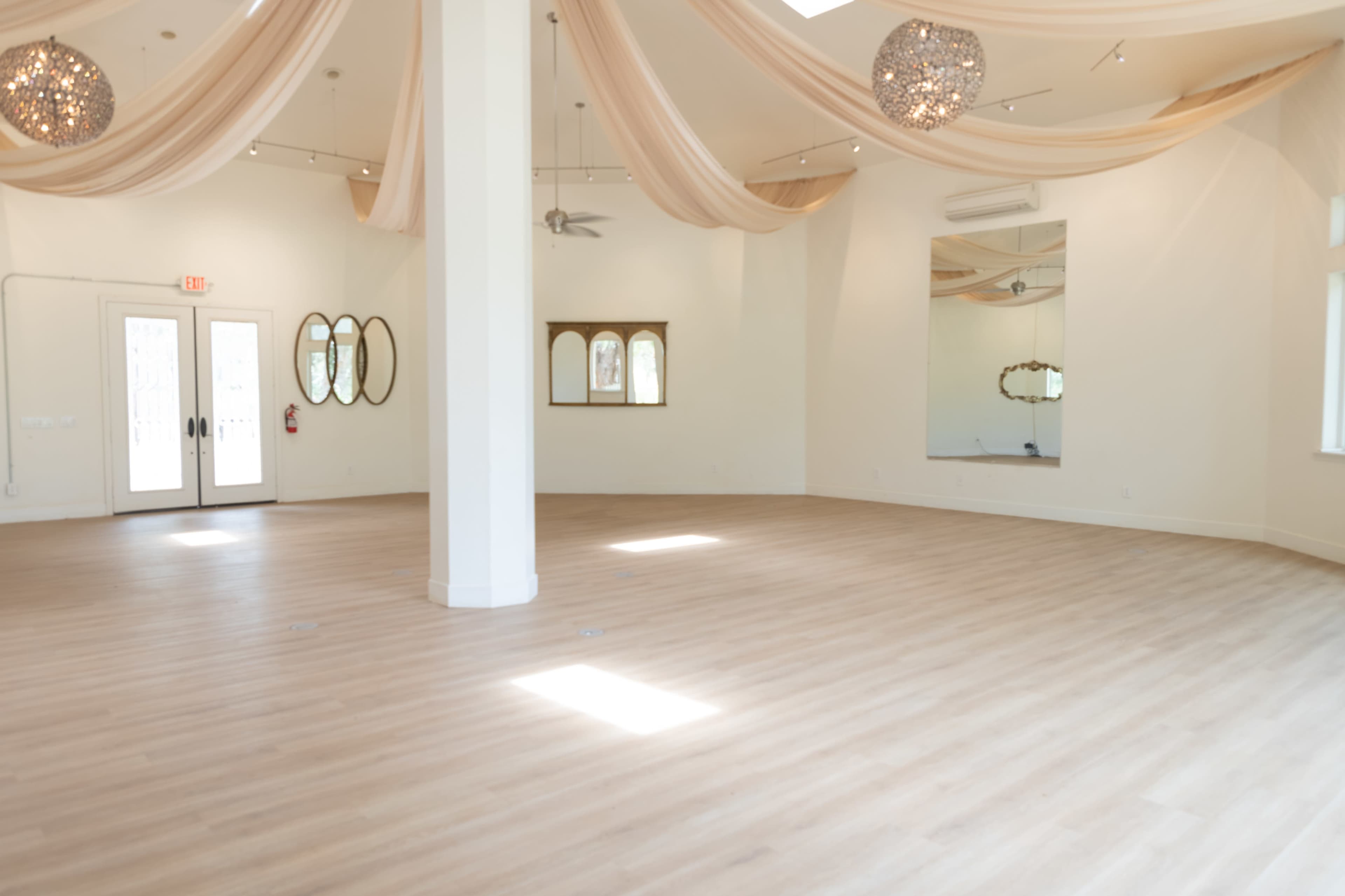 The image shows a spacious and well-lit room with light wood flooring, draped fabric hanging from the ceiling, and large mirrors on the walls.