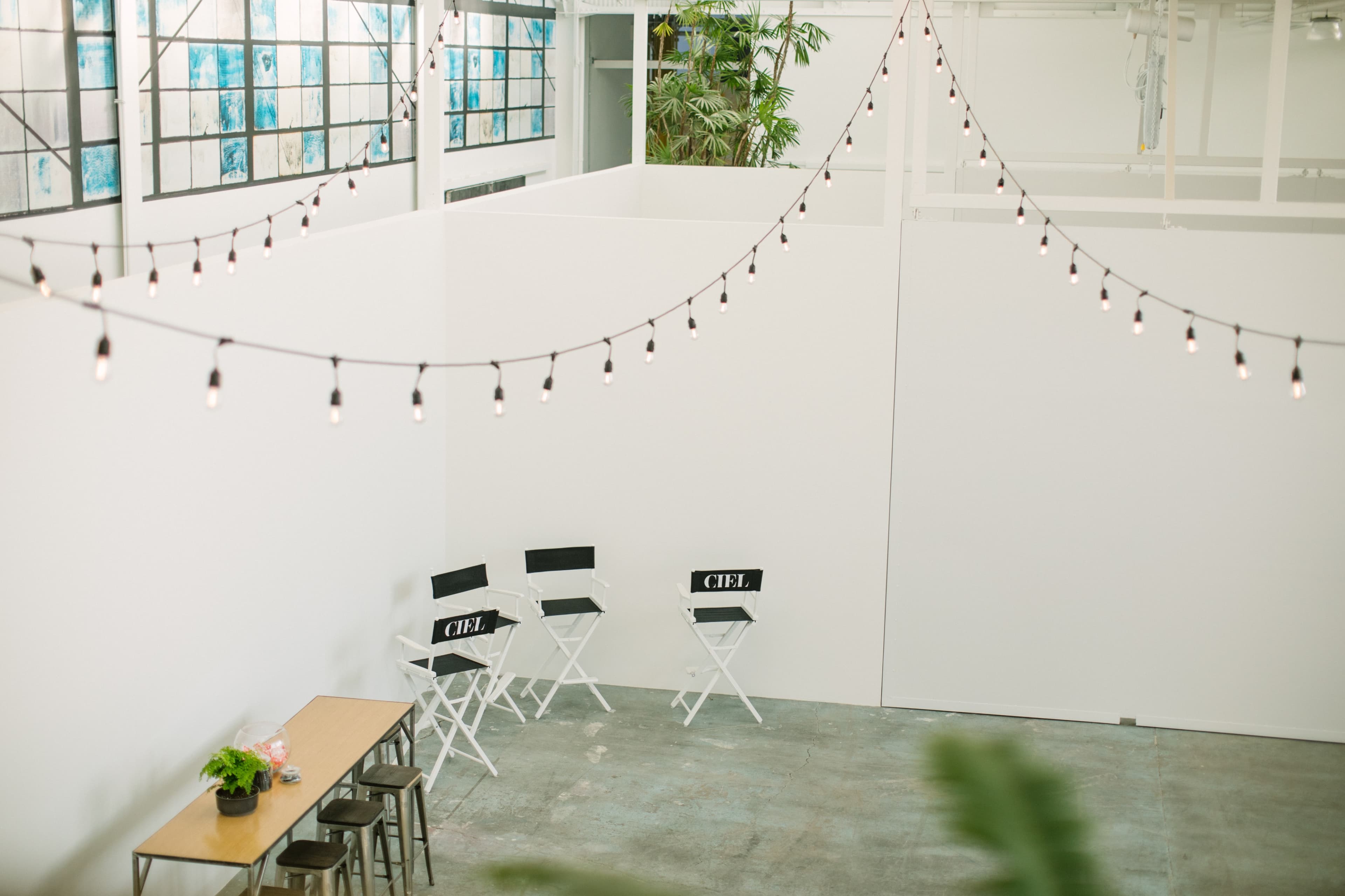 A bright, minimalist interior space with string lights overhead, a few folding chairs labeled "CIEL," and a wooden table.